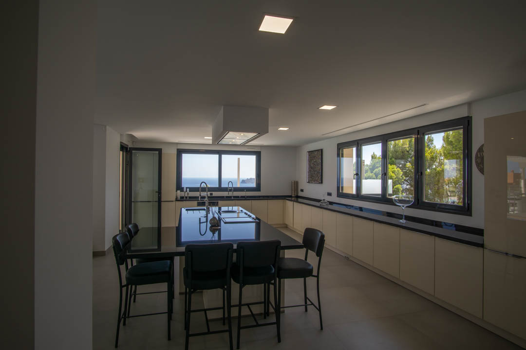 Modern kitchen featuring a large black stone island with seating, a ceiling-mounted extractor, and expansive windows overlooking the Mediterranean Sea and pine forests.