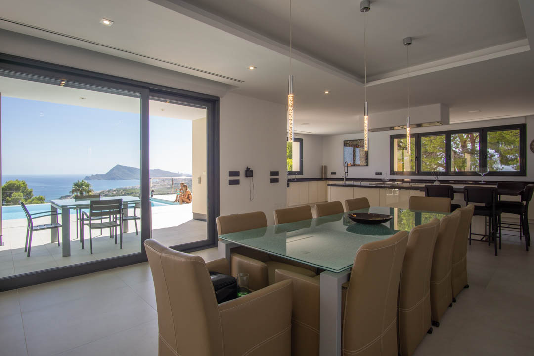 Open-plan dining area with glass table and leather chairs, featuring a modern kitchen and views of the Mediterranean Sea through sliding doors.