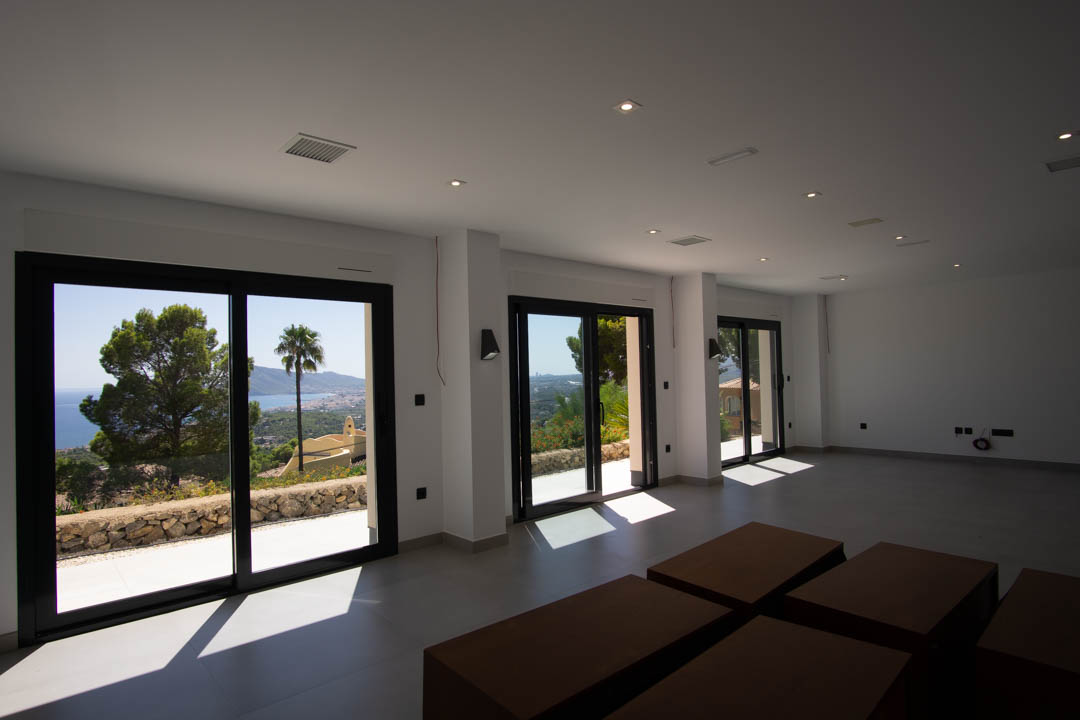 Spacious open-plan living area featuring large black-framed sliding glass doors, recessed ceiling spotlights, and panoramic views of the Mediterranean coastline.