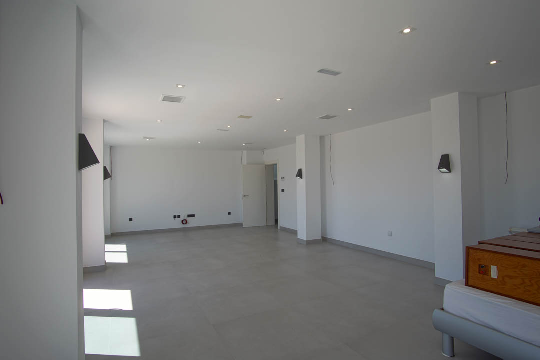 Spacious open-plan room featuring grey tiled flooring, white walls, recessed ceiling spotlights, and contemporary wall-mounted light fixtures.