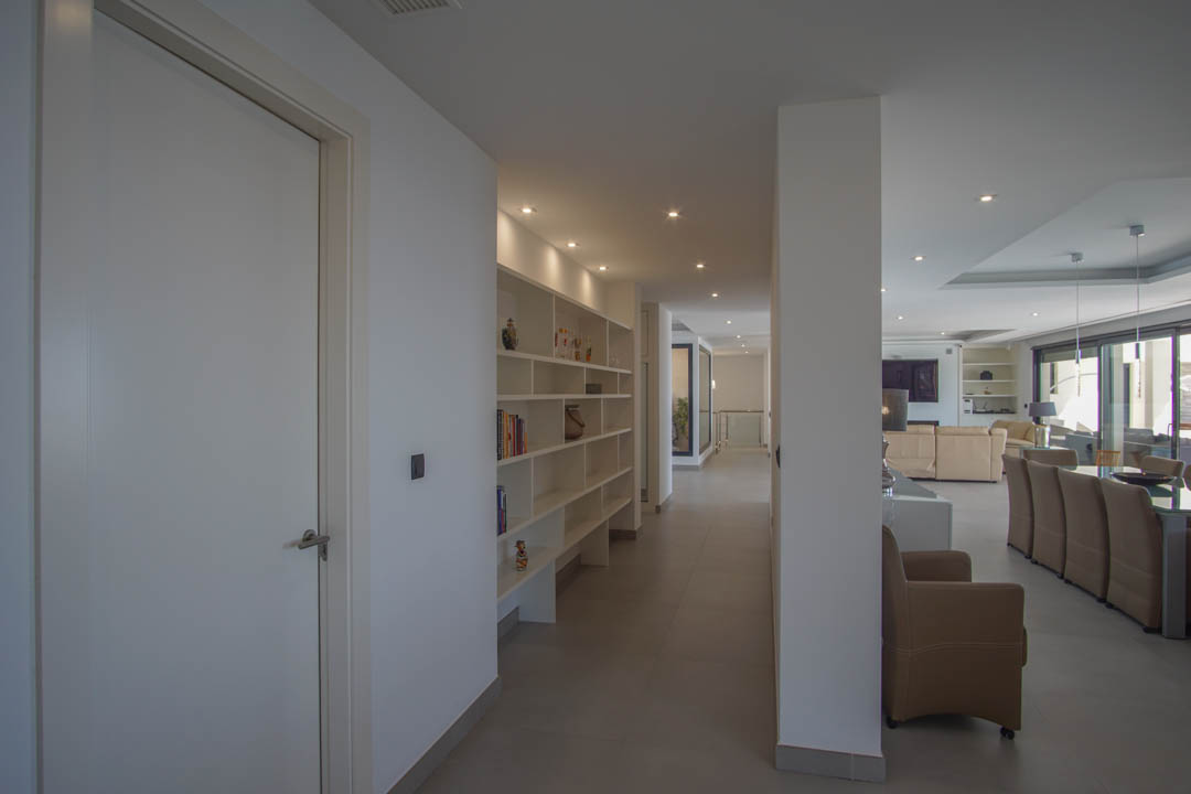 Wide hallway with built-in shelving and recessed lighting, transitioning into an open-plan living and dining area with neutral tones and large windows.