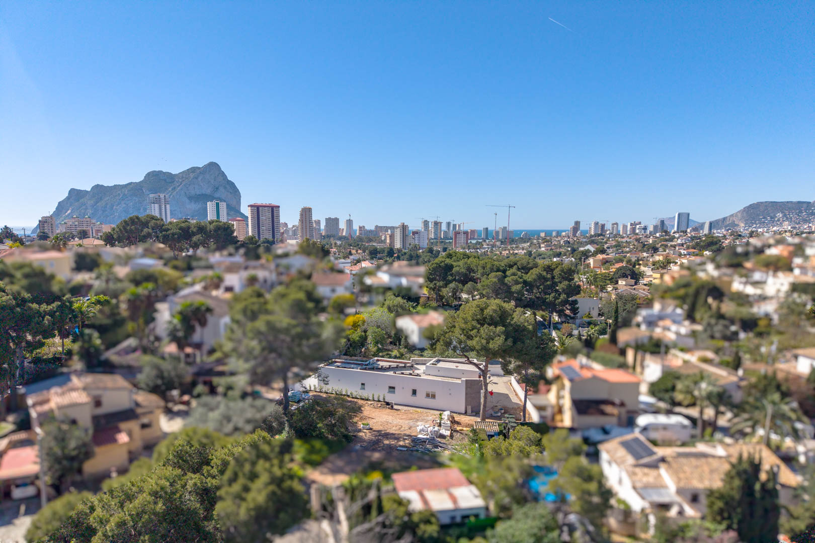 Aerial view of the minimalist villa featuring the Peñón de Ifach and Calpe skyline. The single-story structure is set on a plot awaiting exterior landscaping.