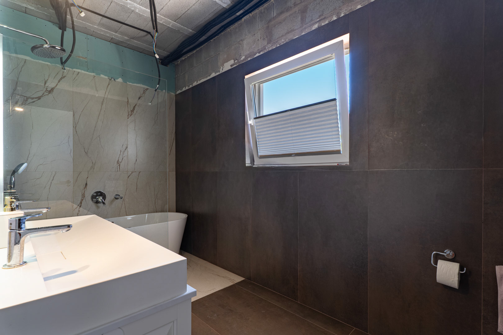Bathroom under completion featuring a double vanity, freestanding bathtub, and marble-effect shower tiling. Large-format dark wall tiles contrast with the light from the tilt-and-turn window.