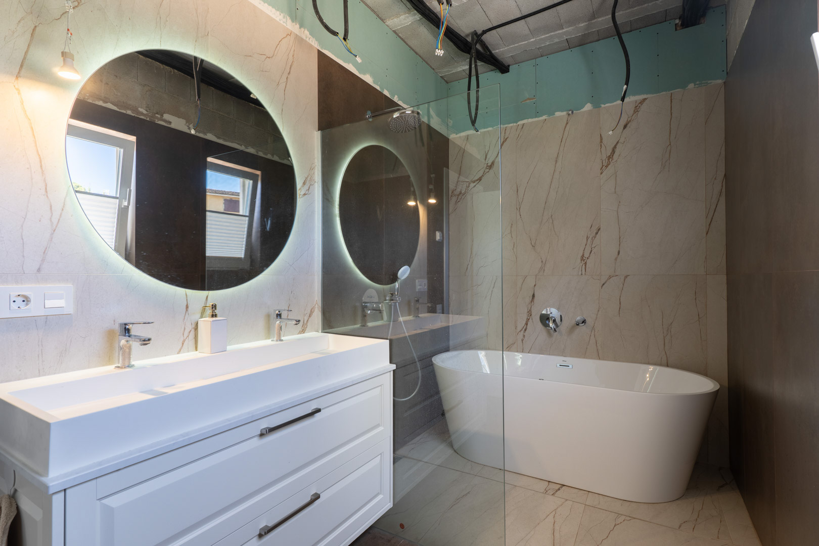 Bathroom under construction featuring a double vanity, backlit circular mirror, and freestanding bathtub against marble-effect porcelain tiling.