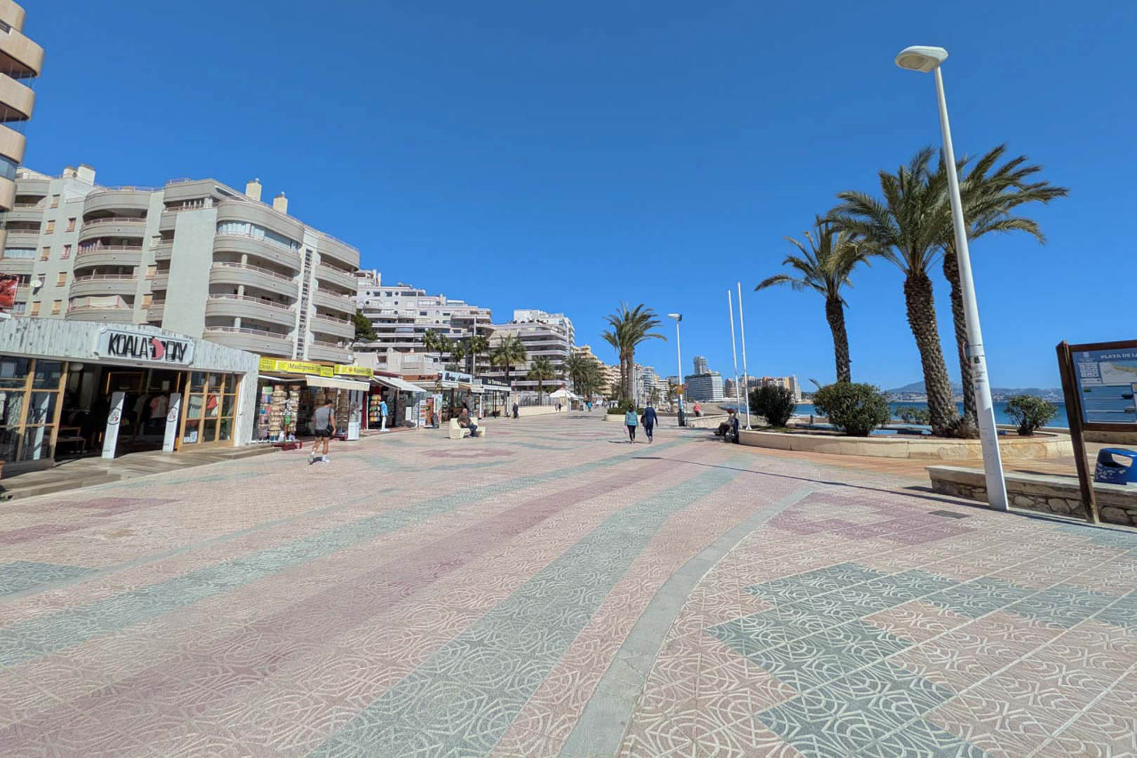 Calpe's beachfront promenade featuring patterned stone paving, local boutiques, palm trees, and multi-story residential buildings under a clear sky.