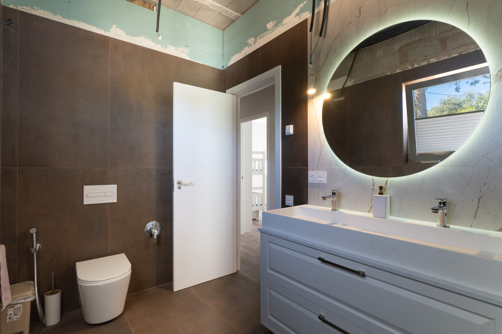 Contemporary bathroom featuring a double vanity, backlit circular mirror, and dark earth-toned tiling. Includes a wall-mounted toilet and white minimalist cabinetry.