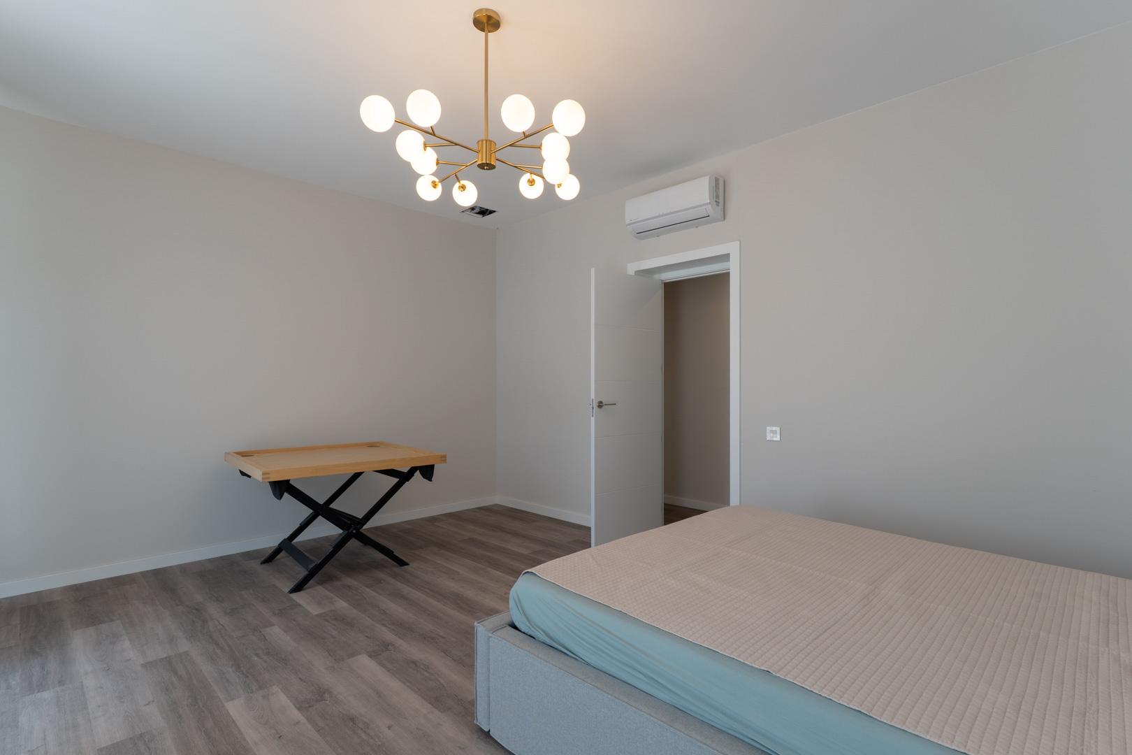 Contemporary bedroom featuring grey wood-effect flooring, neutral walls, and a modern gold chandelier. Equipped with air conditioning and a minimalist white door.