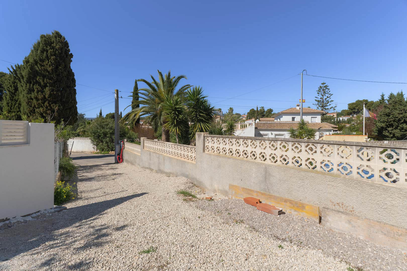 Exterior property access featuring a white gravel driveway and a decorative concrete block perimeter wall, framed by Mediterranean palms and greenery.