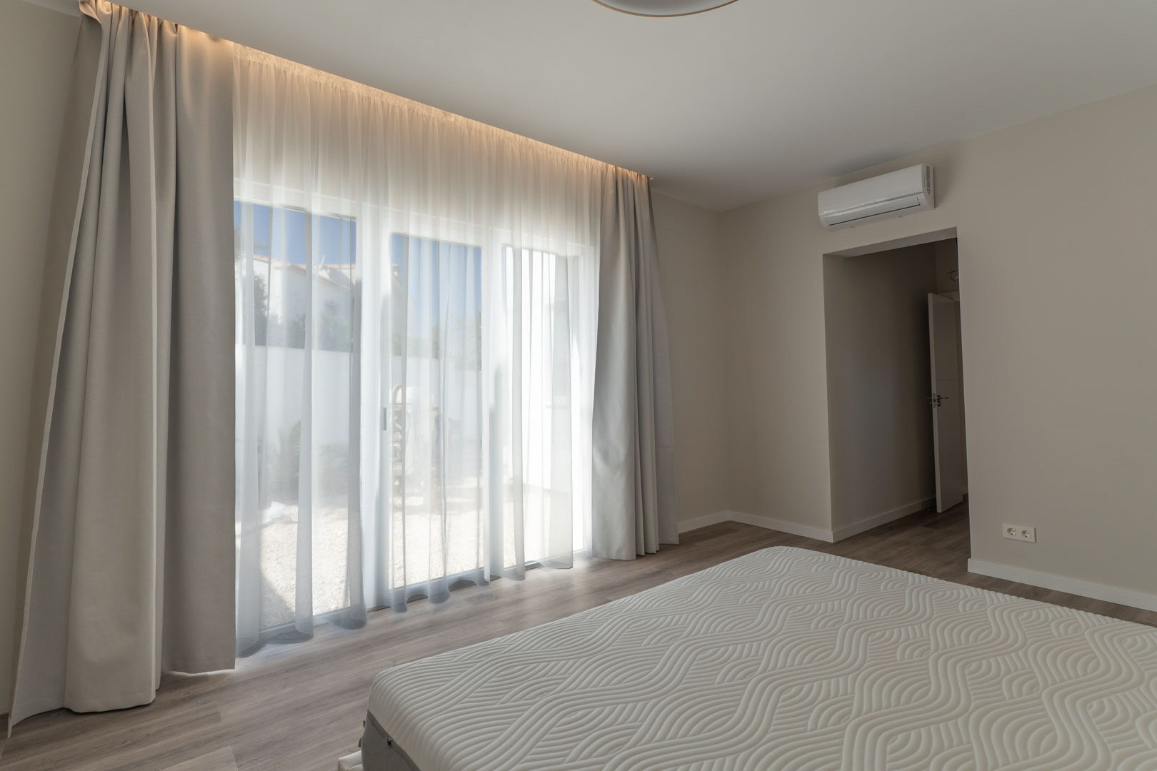 Minimalist bedroom featuring floor-to-ceiling glass doors with integrated LED curtain lighting. Includes split-system air conditioning and light wood-effect flooring.