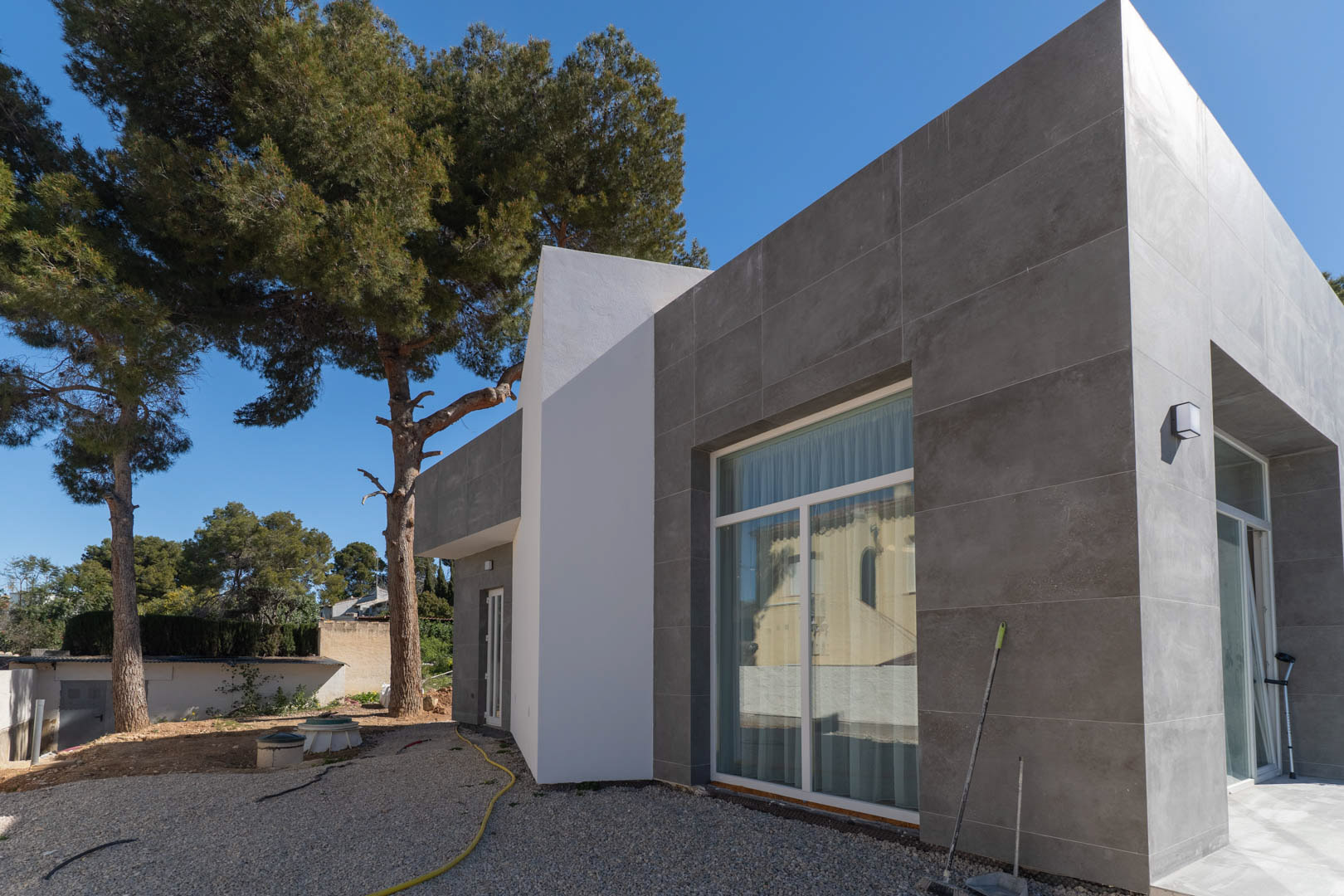 Minimalist exterior featuring grey stone-effect tiling and large floor-to-ceiling windows, set against a backdrop of mature pine trees and unfinished grounds.