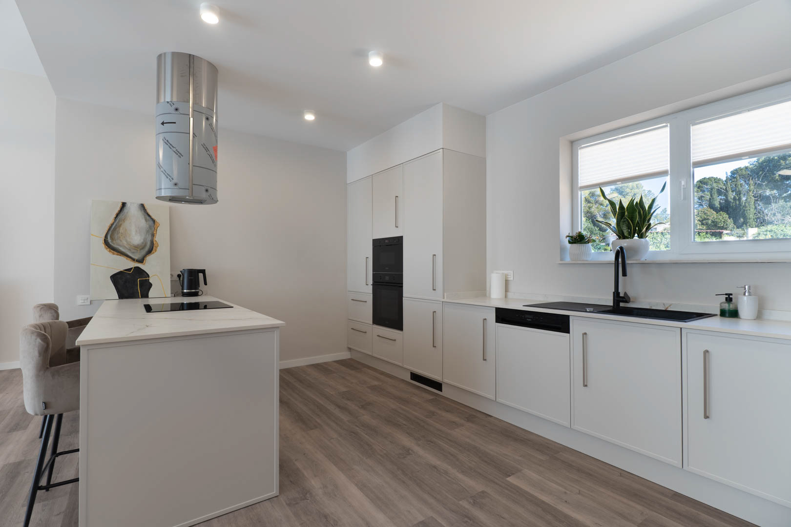 Modern kitchen featuring a central island with induction hob, cylindrical extractor, and integrated appliances. White cabinetry is paired with black fixtures and wood-effect flooring.