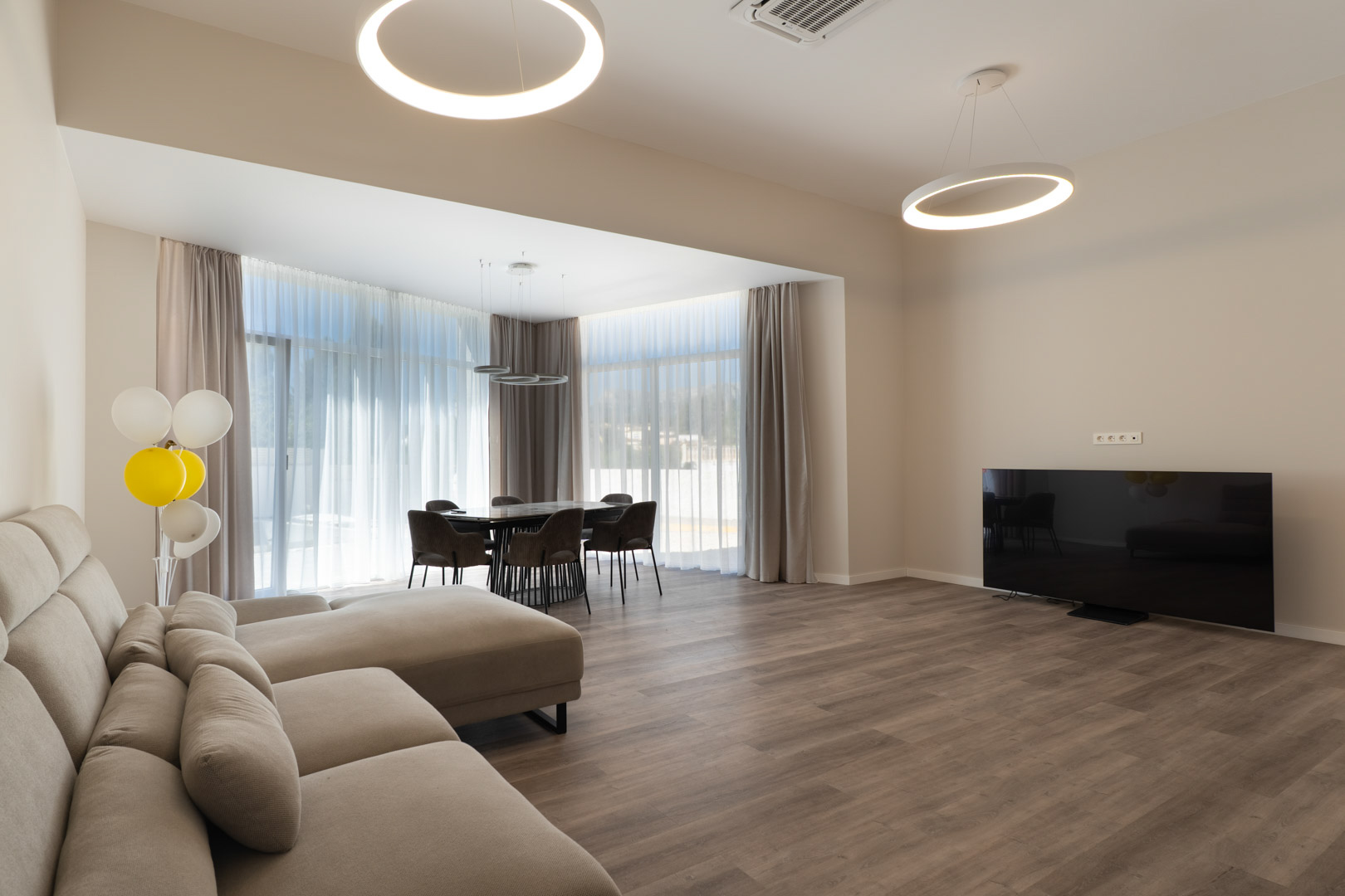 Open-concept living and dining space featuring wood-effect flooring, modern ring pendant lights, and floor-to-ceiling windows with sheer curtains.