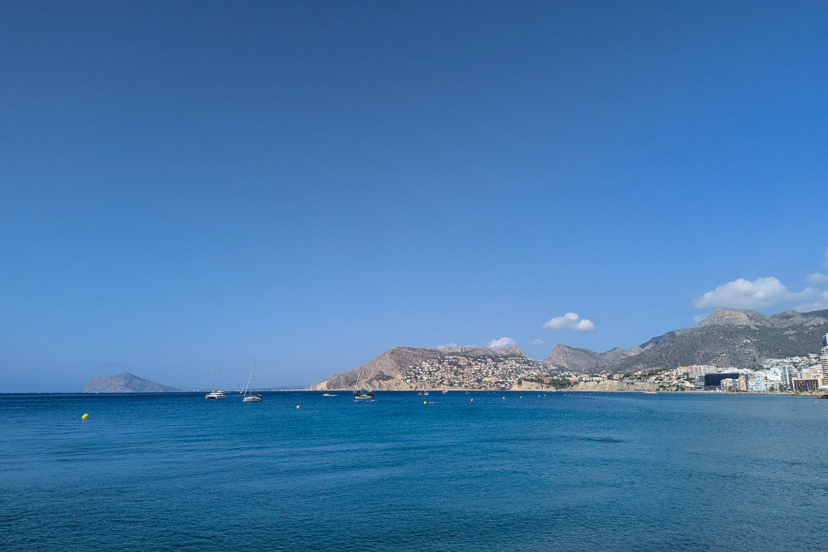 Panoramic view of the Mediterranean Sea off the Calpe coast, featuring sailboats and the mountainous shoreline under a clear blue sky.