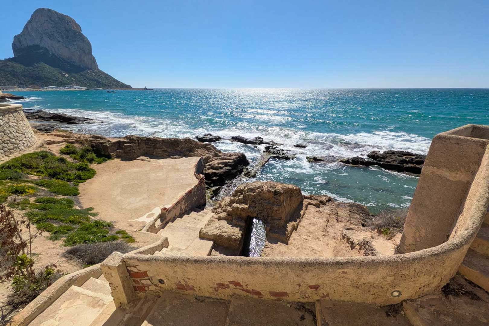 Panoramic views of the Peñón de Ifach from a tiered stone terrace, featuring direct access to a turquoise water cove and the Mediterranean coastline.