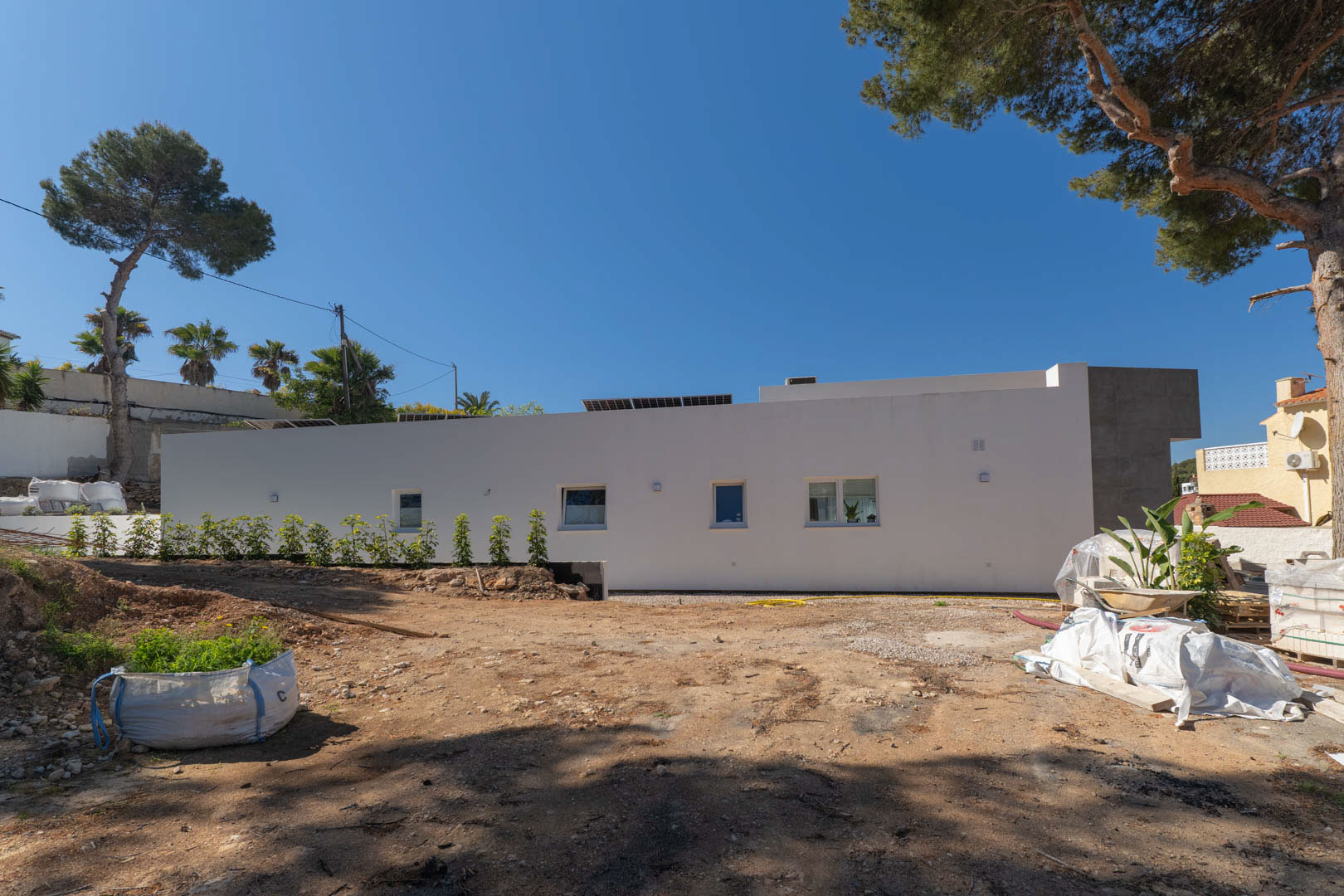 Side facade of the minimalist villa featuring solar panels, showing the exterior grounds currently under preparation and surrounded by mature pine trees.
