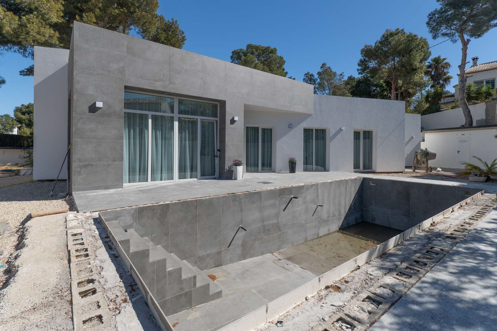 Single-story minimalist villa featuring grey porcelain cladding and an unfinished swimming pool set against a backdrop of mature pine trees.