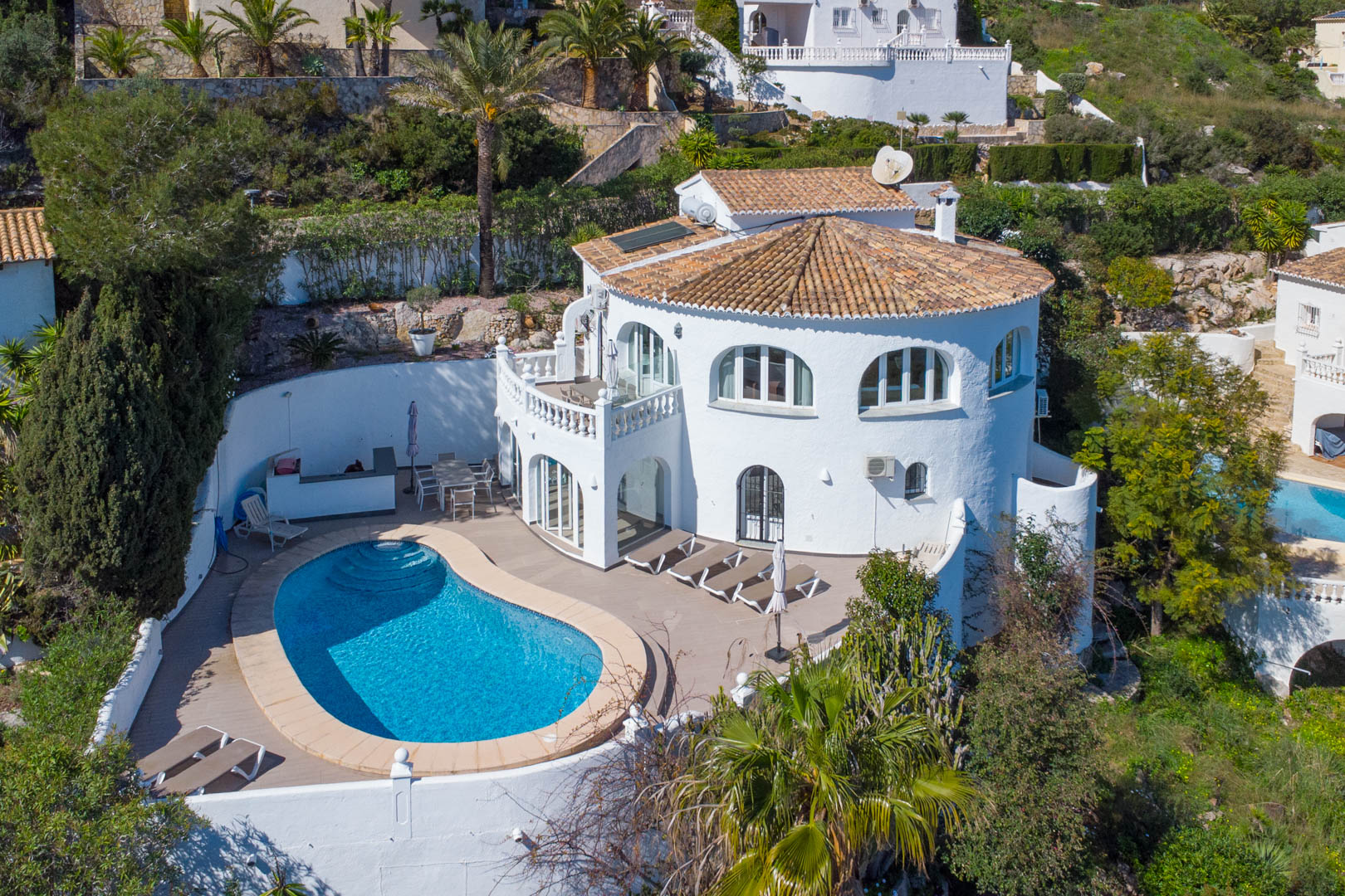Aerial view of the villa featuring a kidney-shaped pool, spacious sun terrace, traditional white facade, and solar panels on the terracotta tile roof.