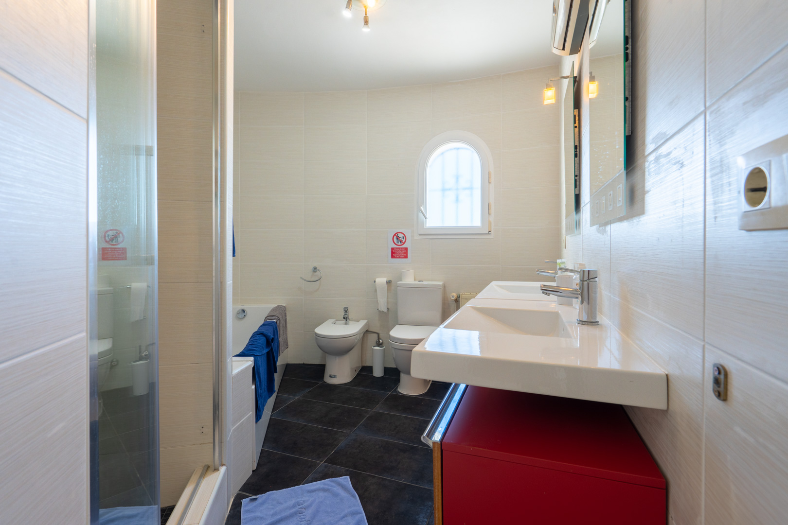 Bathroom featuring a double vanity with a red cabinet, toilet, bidet, and bathtub. The space includes light wall tiling, dark flooring, and an arched window.