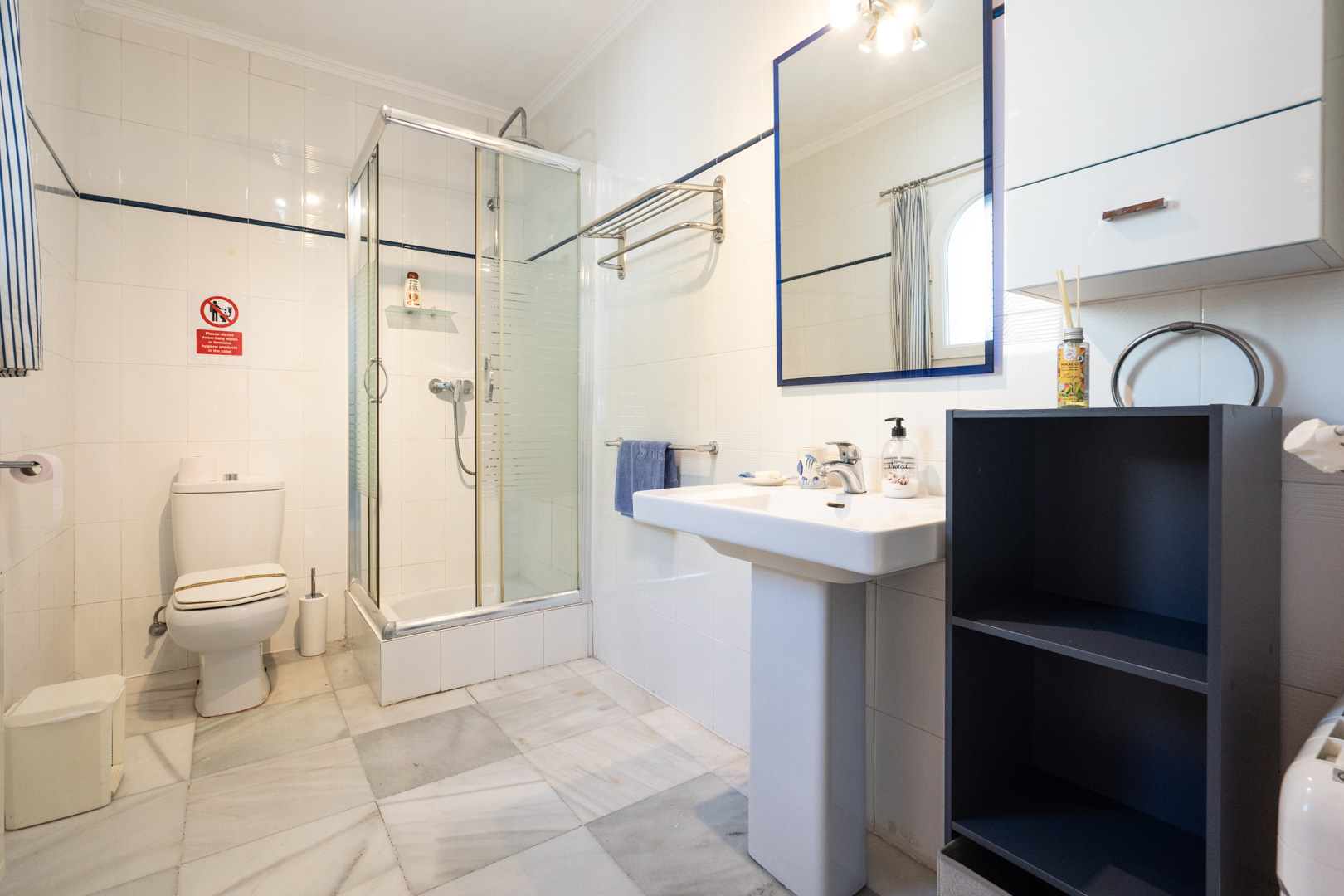 Bathroom featuring white wall tiles with blue accents, marble flooring, a glass corner shower, and a pedestal sink with a blue-framed mirror.