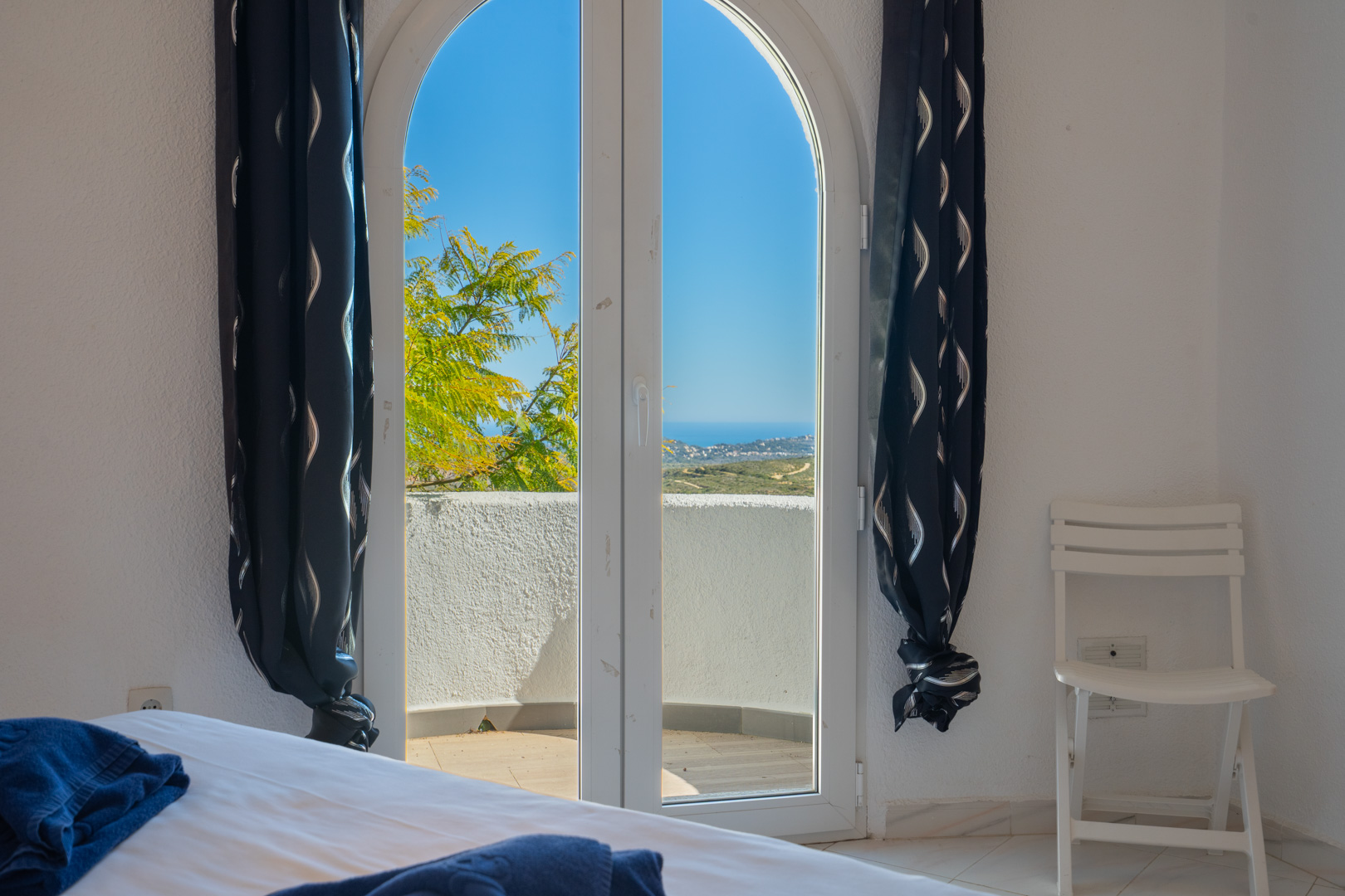 Bedroom featuring an arched glass door opening onto a private balcony with panoramic sea views. The room includes white walls and dark patterned curtains.