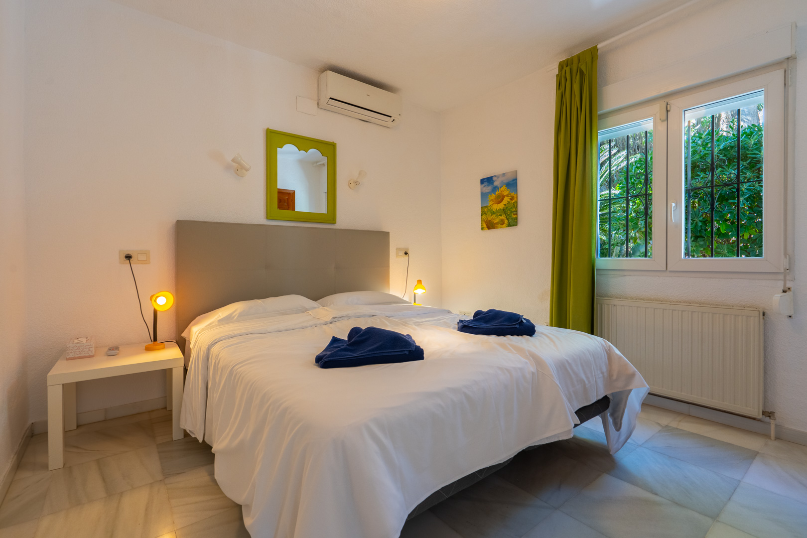 Bedroom with marble flooring, air conditioning, and heating. It features a window with security bars, green curtains, and a matching decorative mirror frame.
