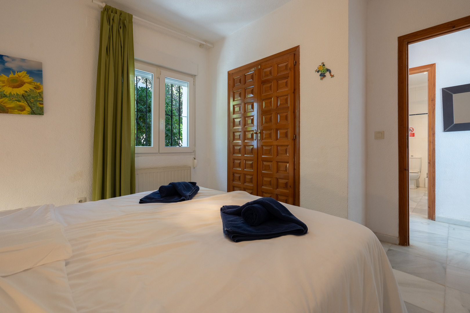 Bright bedroom featuring a carved wooden built-in wardrobe, green curtains, and a doorway leading to an ensuite bathroom with marble tiles.