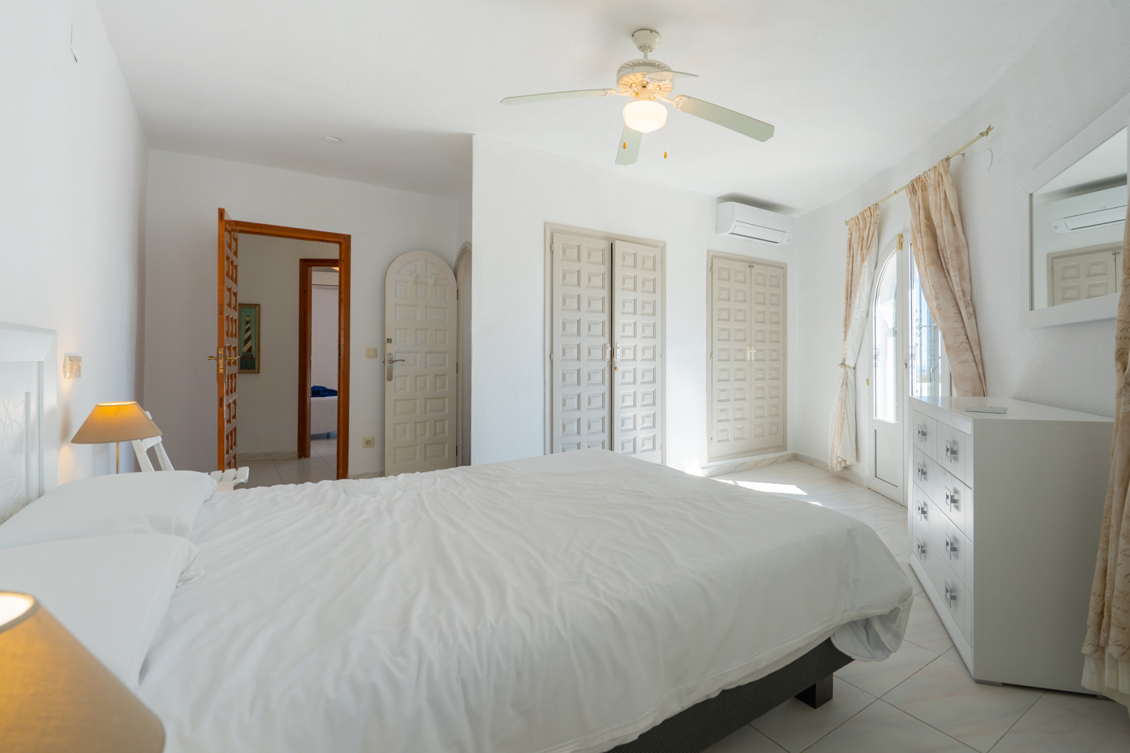 Bright bedroom featuring traditional built-in wardrobes, air conditioning, a ceiling fan, and direct outdoor access through an arched glass door.