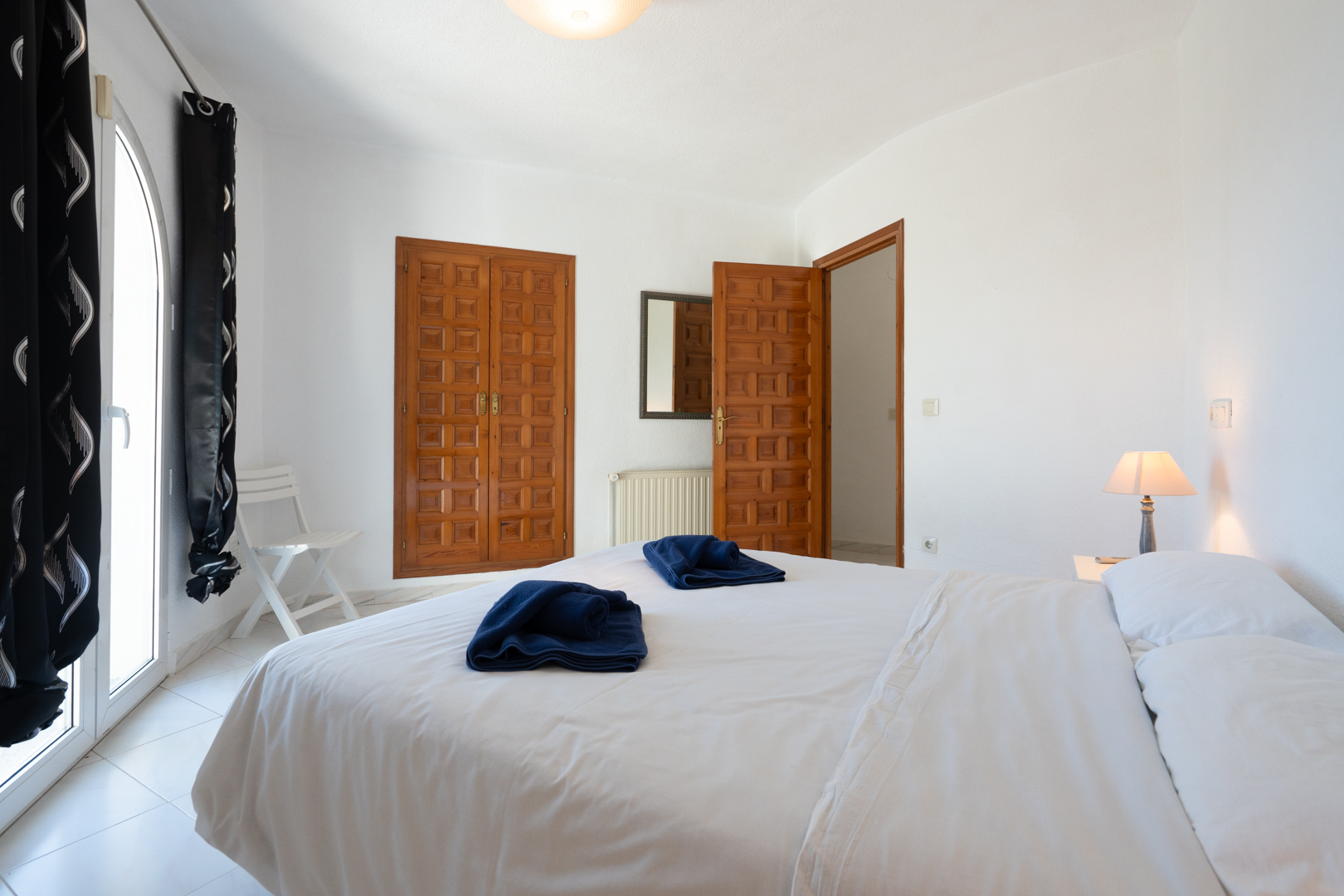 Bright bedroom featuring traditional wood-paneled built-in wardrobes, an arched glass door for natural light, and clean white walls with tiled flooring.