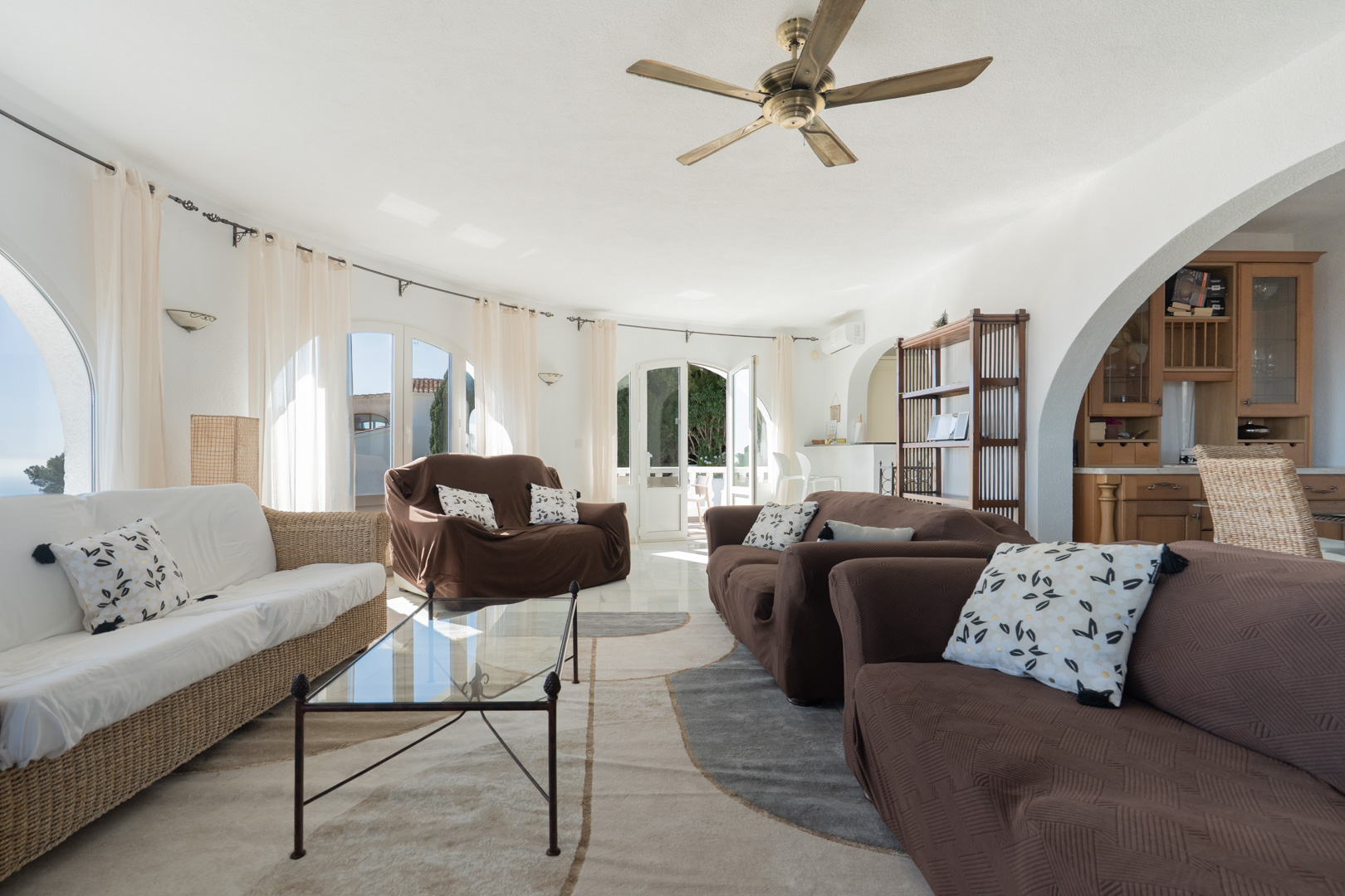 Bright living room featuring a curved wall with arched windows and terrace access. It includes a wooden ceiling fan, tiled floors, and an arched opening to the kitchen.
