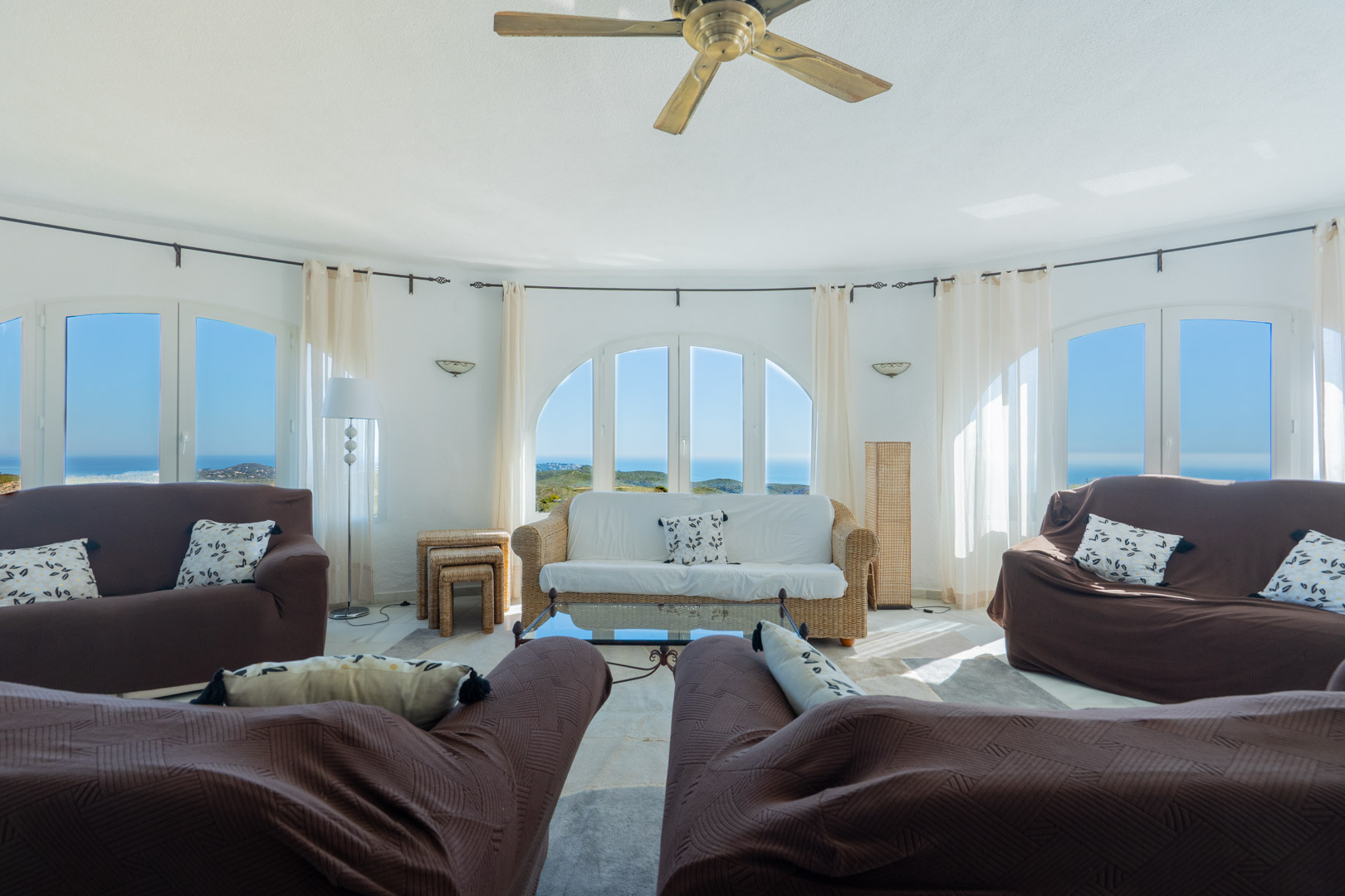 Circular living room featuring arched windows with panoramic Mediterranean views. The space includes a ceiling fan, wicker furniture, and natural light.