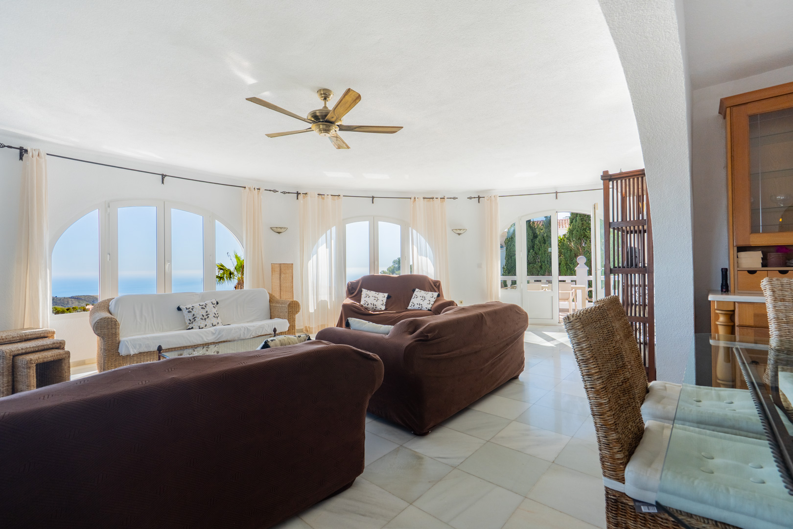 Circular living room featuring arched windows with sea views, light marble tiling, a wooden ceiling fan, and traditional wicker furniture accents.
