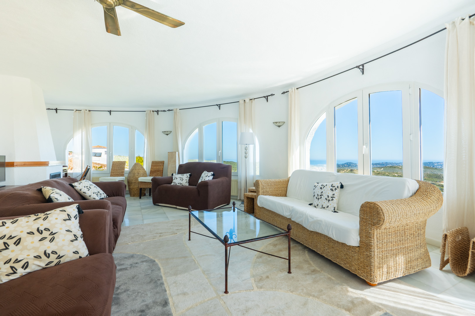 Circular living room featuring large arched windows with sea views, a mix of wicker and fabric seating, and a wooden ceiling fan.