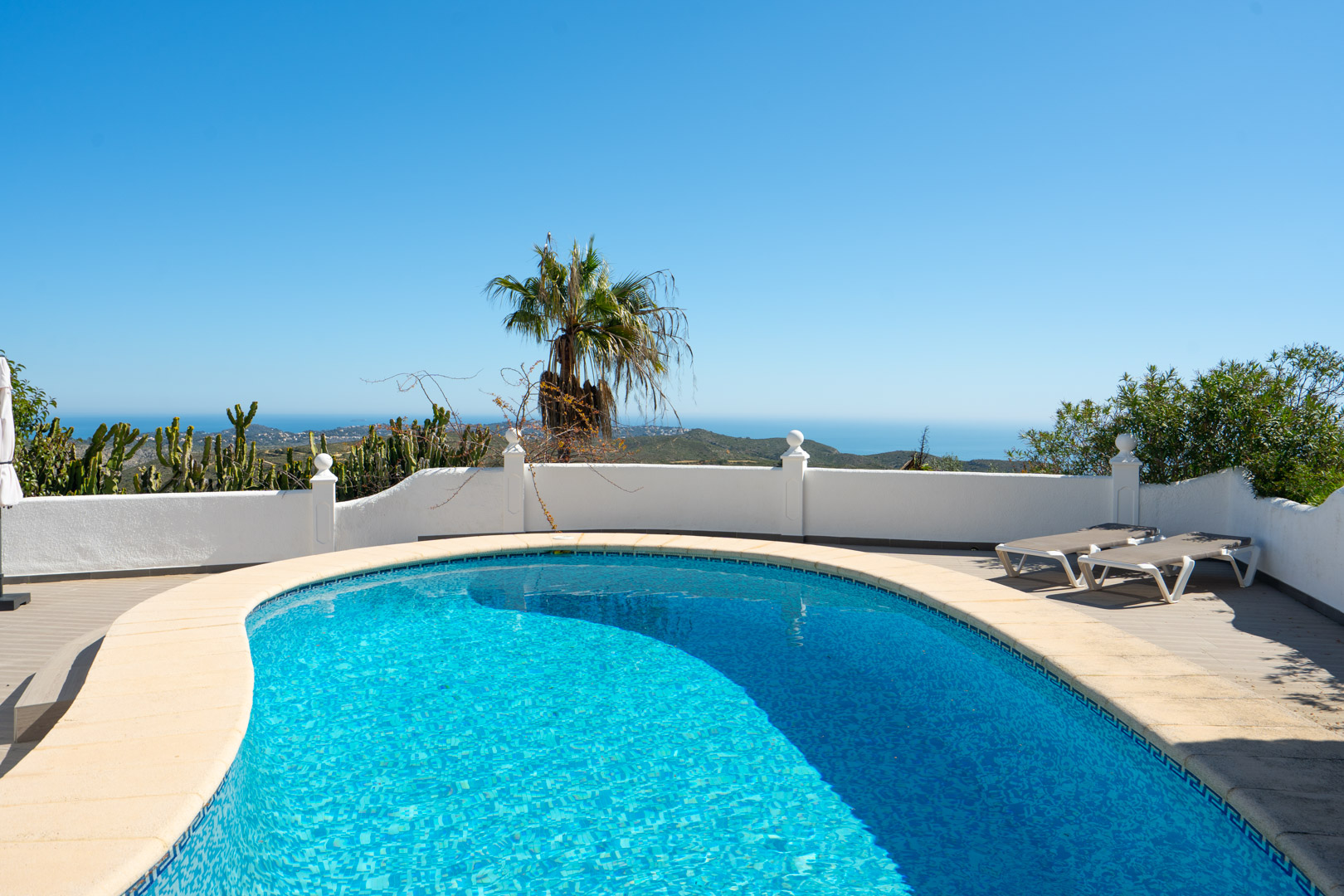 Curved swimming pool overlooking the Mediterranean Sea, featuring a stone-bordered sun terrace, white perimeter walls, and panoramic coastal views.