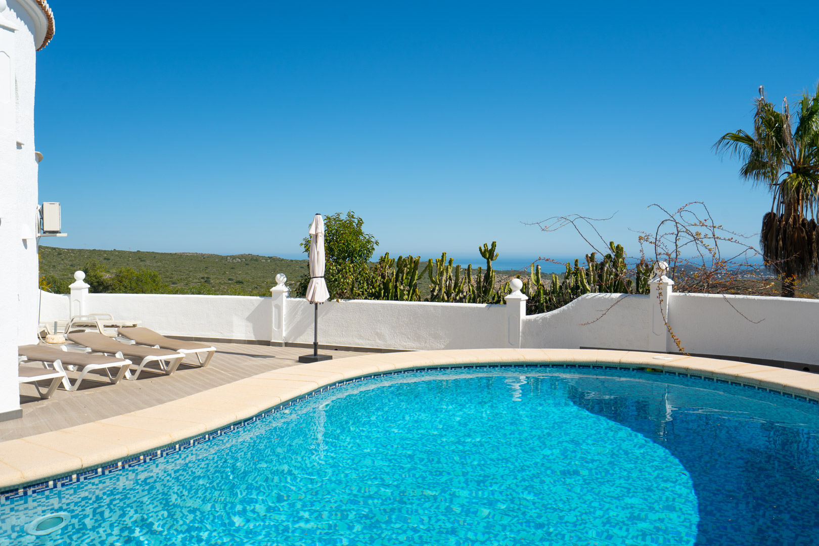 Curved swimming pool surrounded by a sun terrace with loungers, featuring panoramic views of the Mediterranean Sea and the Granadella natural park.