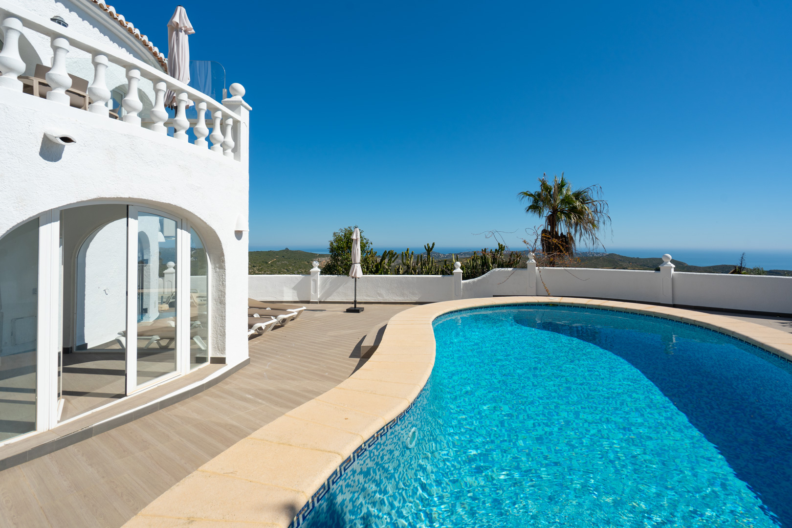 Curved swimming pool with blue mosaic tiling and a spacious wood-effect sun terrace, offering panoramic Mediterranean views next to the arched glass naya.