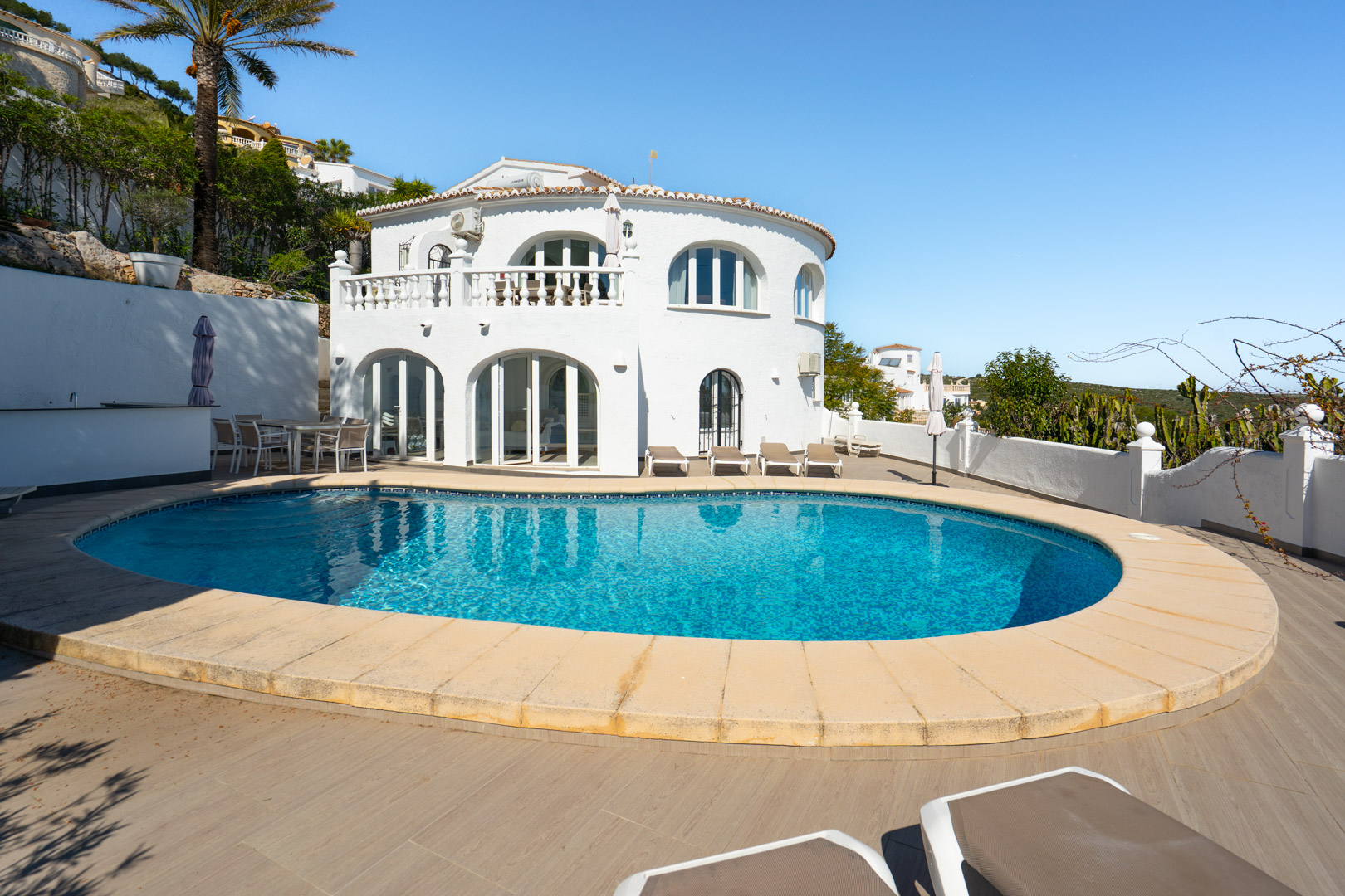 Exterior view featuring a curved swimming pool, a spacious wood-effect tiled terrace with sun loungers, and a white villa with arched windows and a classic balustrade.