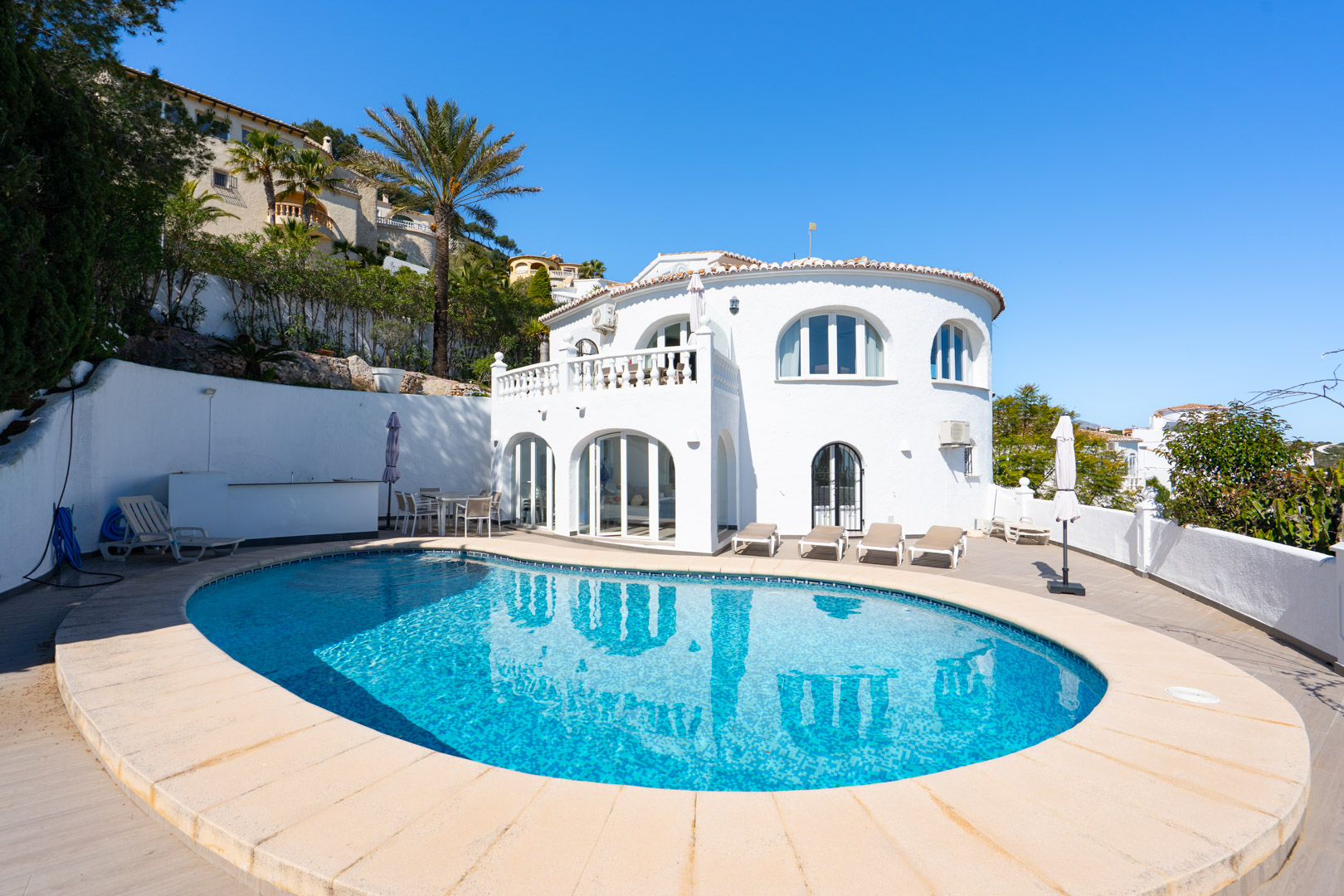 Exterior view featuring an oval swimming pool, a spacious tiled sun terrace, and a traditional white villa with arched windows and a glazed naya.