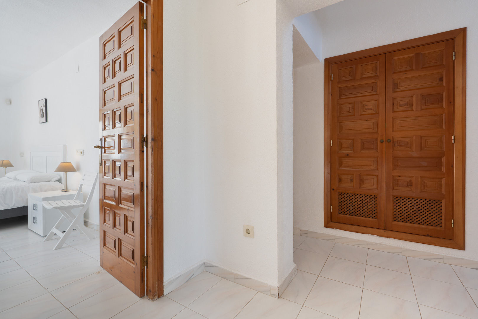Hallway featuring traditional carved wooden doors and built-in wardrobes, leading into a bright bedroom with white tiled flooring and neutral walls.