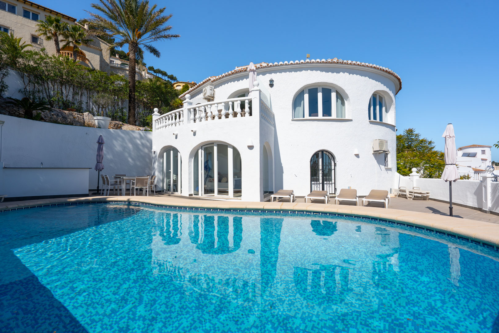 Mediterranean villa exterior featuring a private pool, sun terrace, and glass-enclosed naya. The white facade includes traditional arched windows and balustrades.
