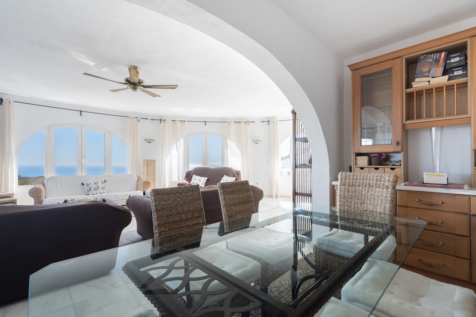 Open-plan living and dining area featuring a glass table, wicker seating, and built-in wooden cabinetry. Arched windows provide natural light and sea views.