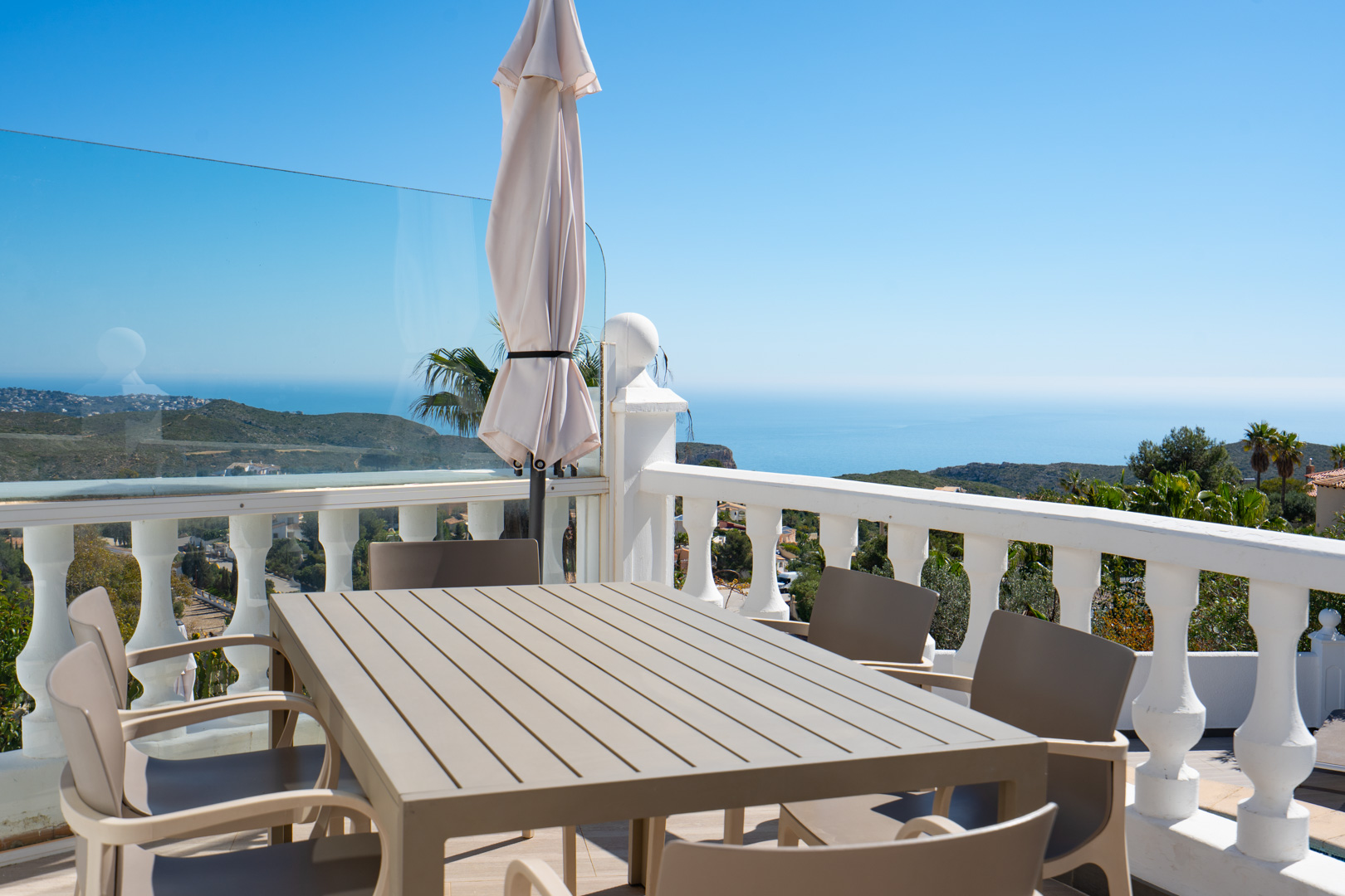 Outdoor dining terrace featuring a slatted table and chairs, enclosed by white balustrades and glass panels with panoramic views of the Mediterranean Sea.
