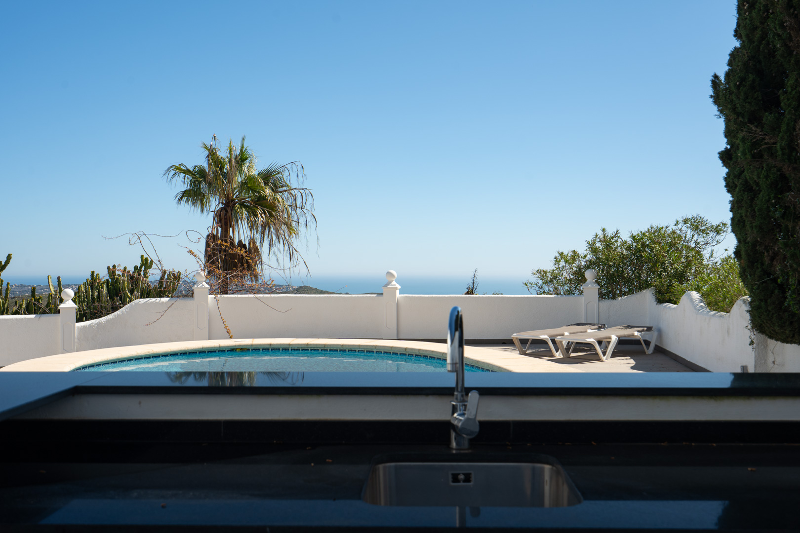 Outdoor kitchen area featuring a dark countertop and sink, overlooking the curved pool and sun terrace with panoramic sea views.