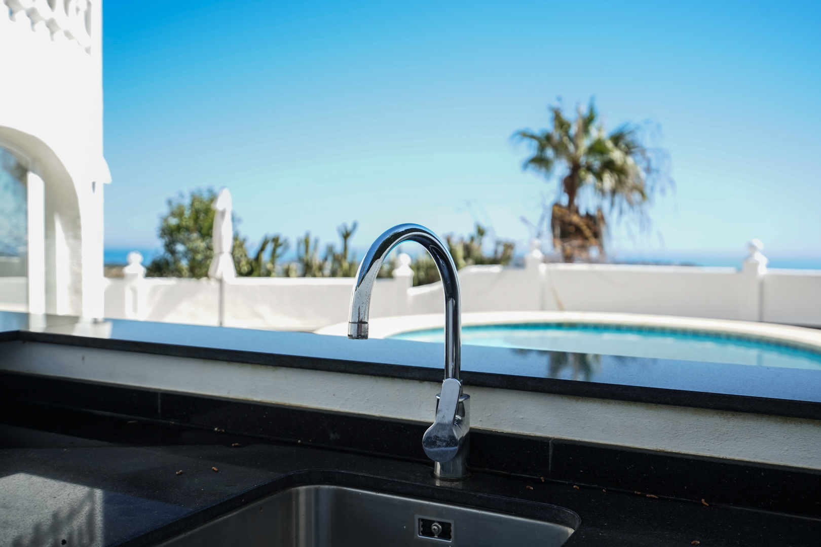 Outdoor kitchen area with a black granite countertop and stainless steel sink, overlooking the private swimming pool and palm trees.