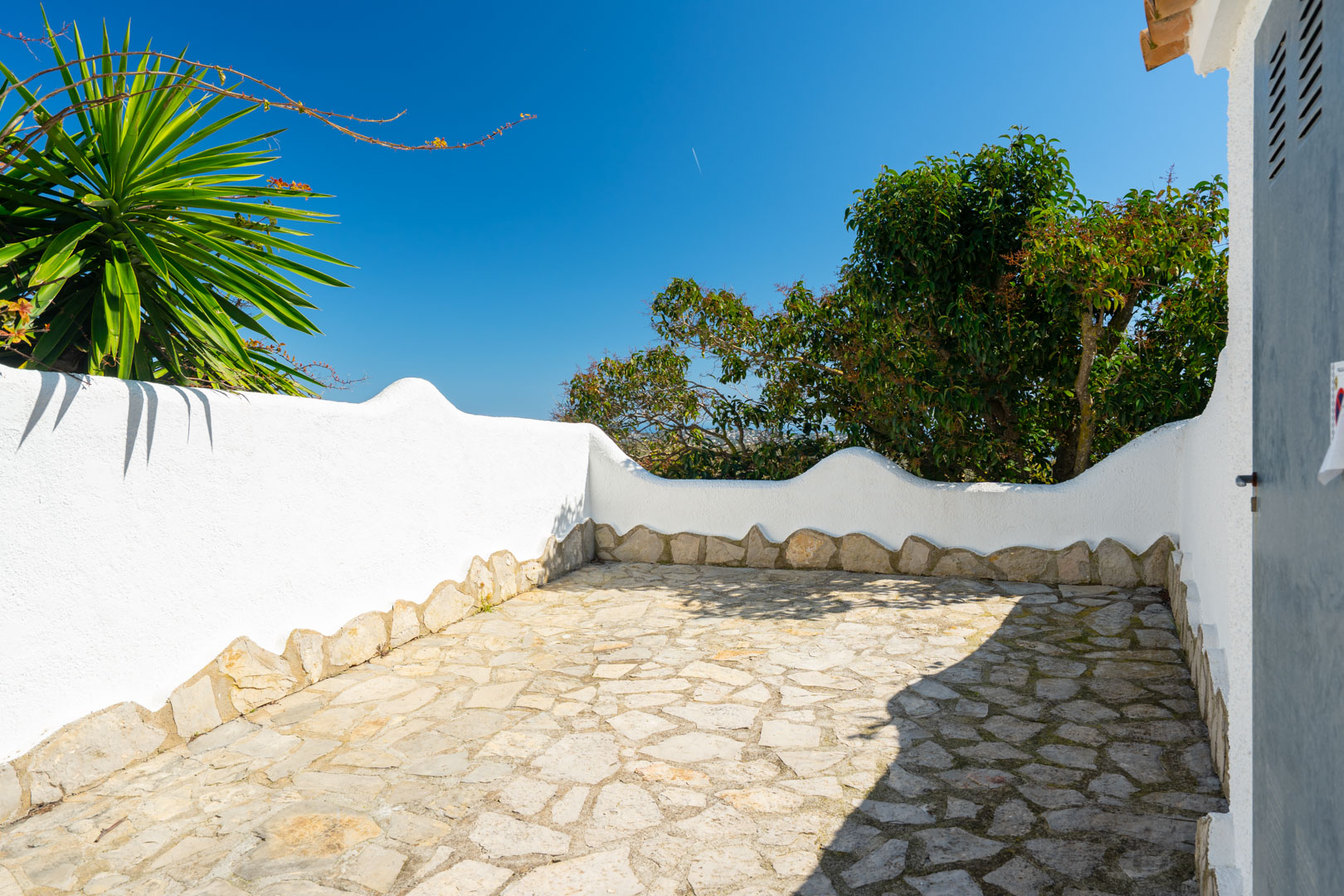 Outdoor terrace featuring irregular natural stone paving and white-washed walls with organic wavy silhouettes, set against Mediterranean greenery.