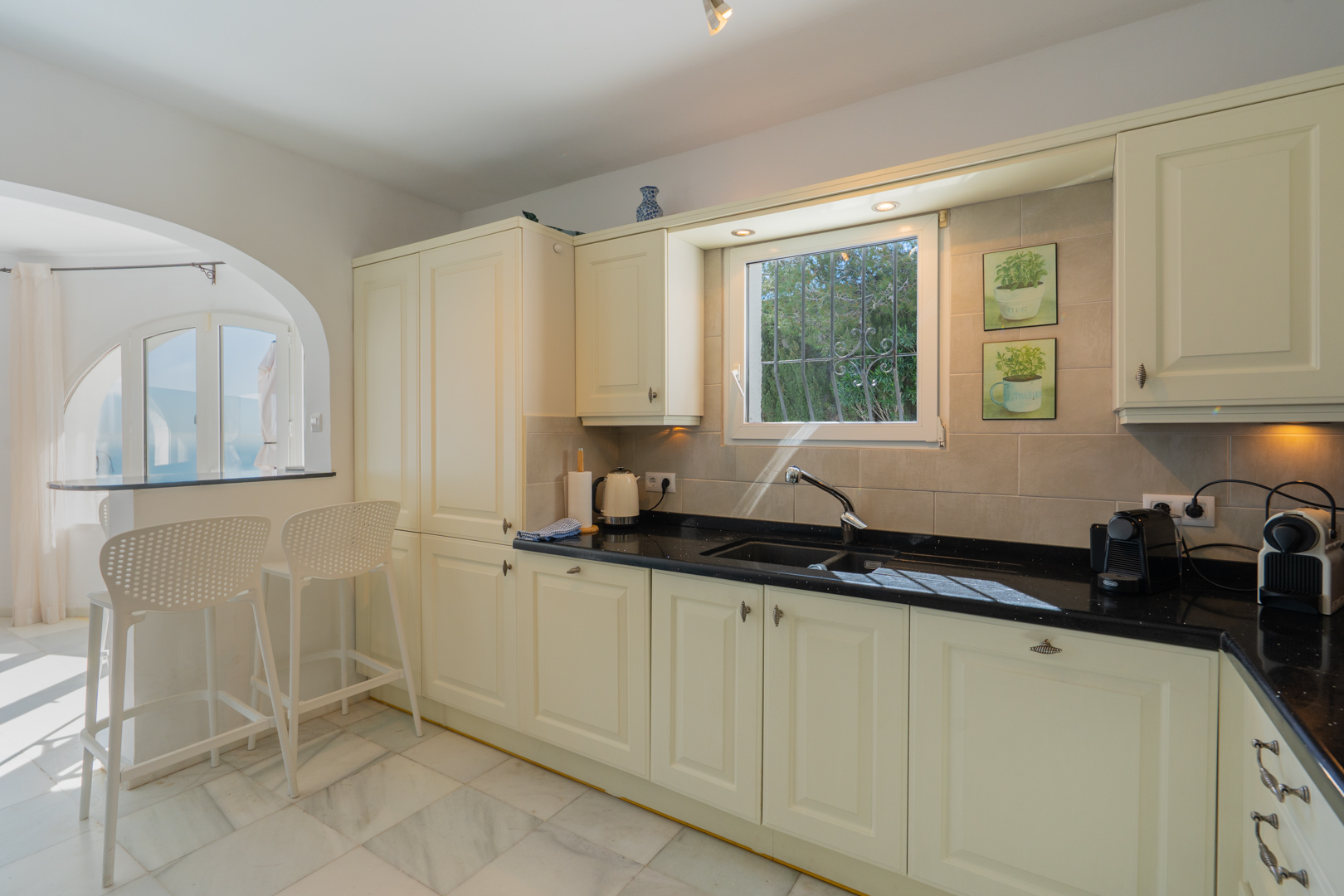 Traditional kitchen featuring cream cabinetry, dark stone countertops, and marble flooring. An arched opening connects the space to a breakfast bar with white stools.