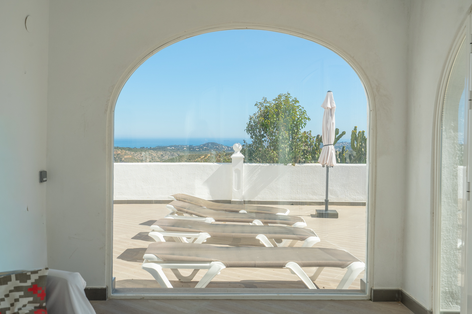 View from the glazed naya through a large archway, showing the sun terrace with loungers and panoramic Mediterranean Sea views.