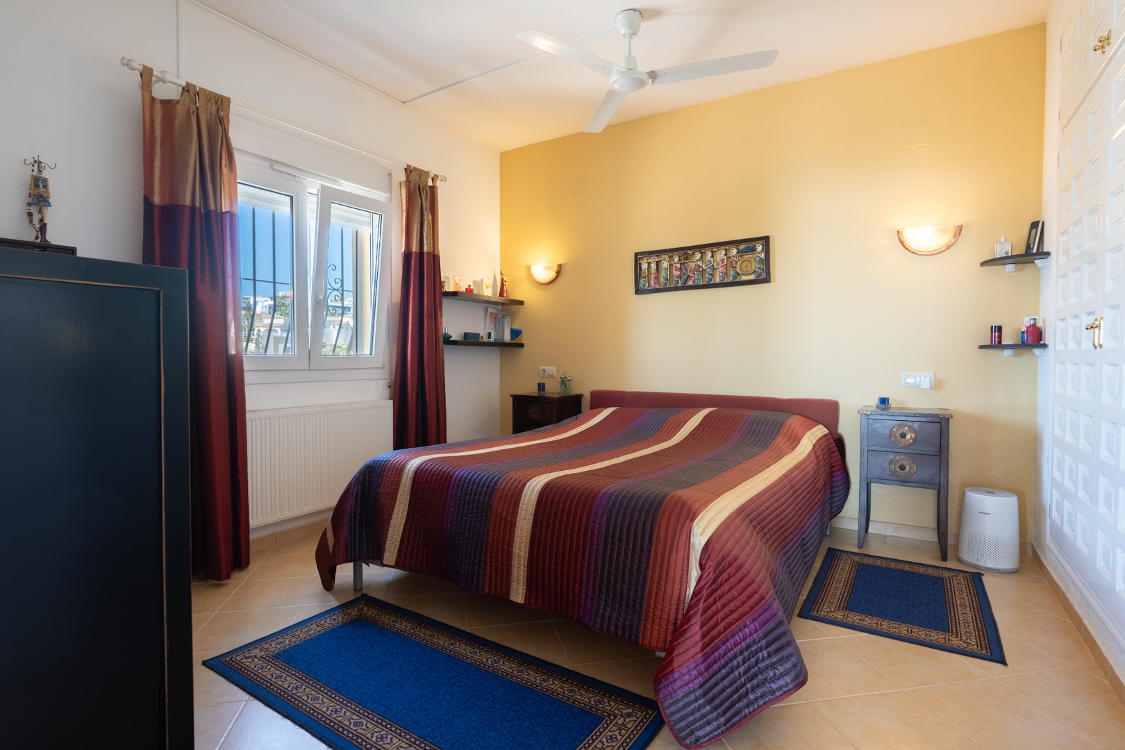 Bedroom featuring a double bed, yellow accent wall, ceiling fan, and white built-in wardrobes. It includes radiator heating and ceramic tiled flooring.