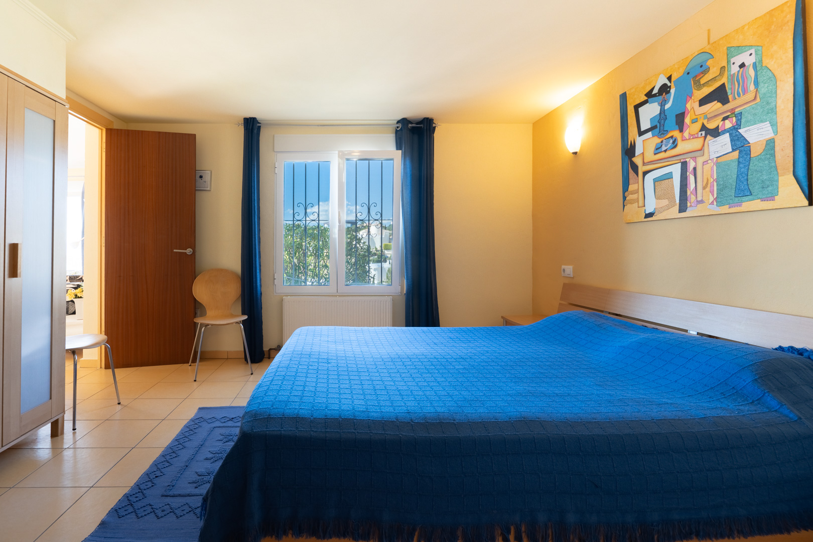 Bedroom with a double bed, blue linens, wooden wardrobe, and tiled flooring. A window with security bars provides natural light and garden views.