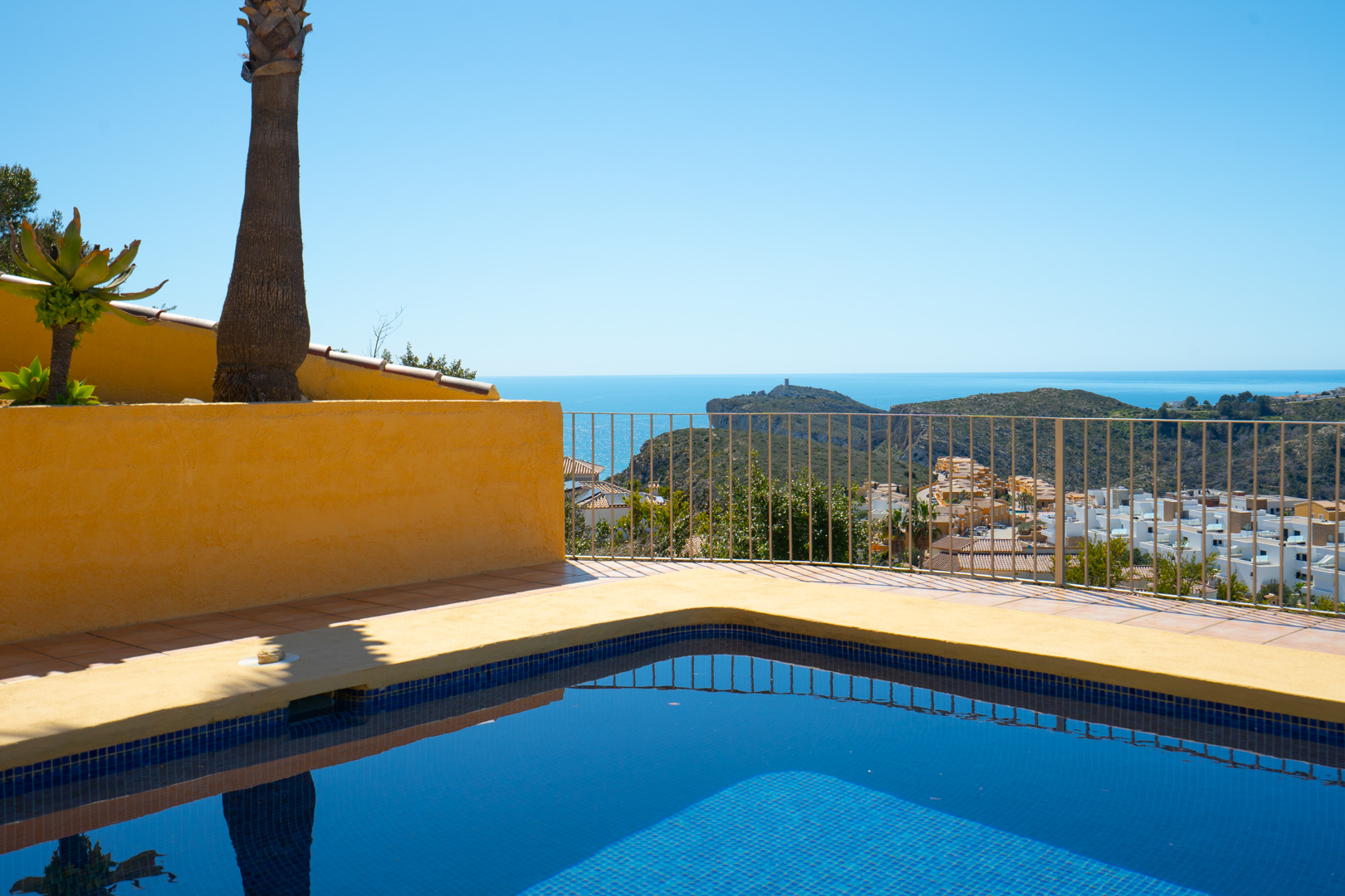 Blue mosaic pool and sun terrace featuring panoramic views of the Mediterranean Sea and the Cap d'Or headland, framed by a classic metal railing.