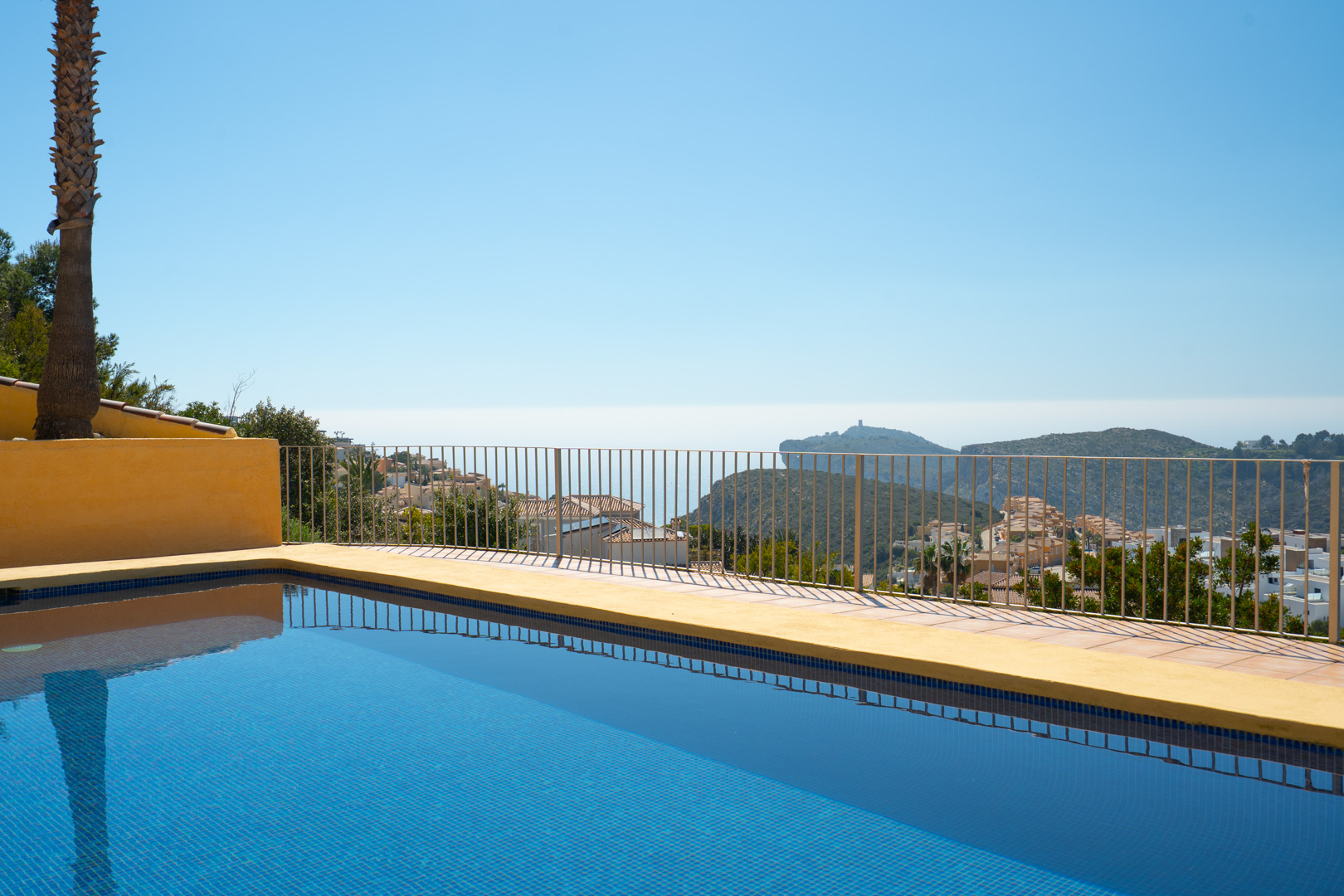 Blue tiled swimming pool featuring panoramic views of the Mediterranean and Cap d’Or, surrounded by a sun terrace with a metal safety railing.
